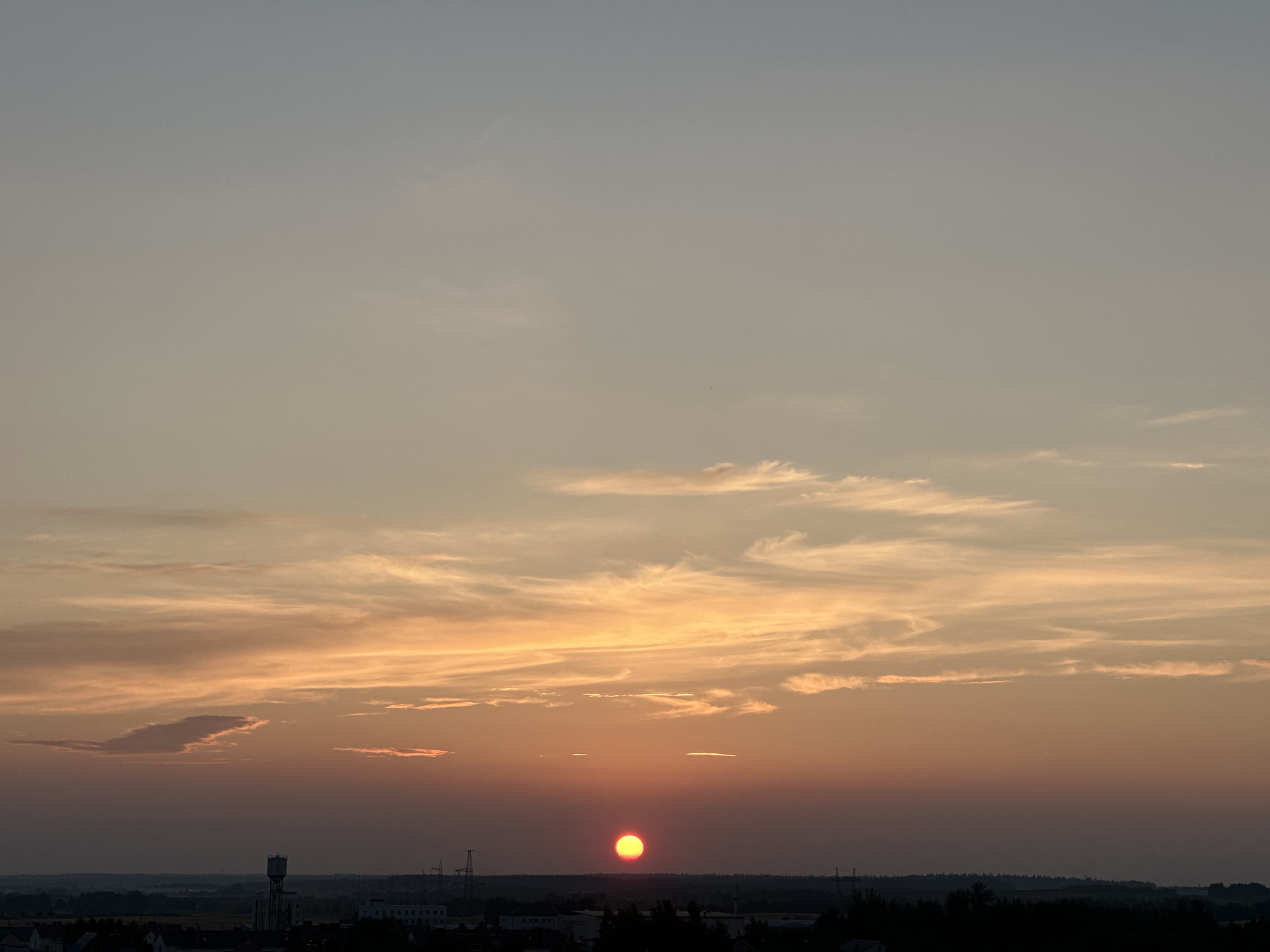 This is a wide shot of a sunset over a rural landscape near the city of Shchomyslitsky Sel'skiy Sovet, Belarus. The sky is filled with soft orange and yellow hues, with wispy clouds scattered across the horizon. A bright red sun is visible near the center of the frame, just above the dark silhouette of trees and buildings that mark the edge of the city. A tall water tower is prominently featured on the left side of the city skyline. The overall scene is peaceful and serene, capturing the beauty of a sunset over a European landscape.