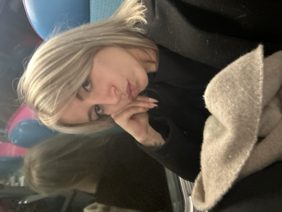 A blonde woman rests her head on her hand. She wears a black jacket. A beige scarf is visible. Her fingernails are manicured. A reflection of her face is visible in the lower-left corner. Blue and pink balloons are in the background, partially obscured. She is looking at the camera.