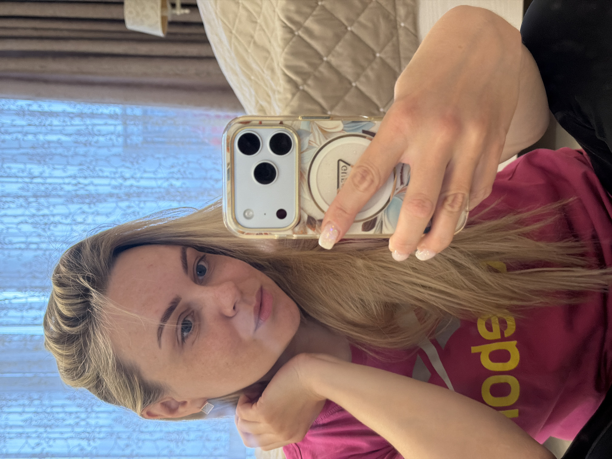A young woman with long, blonde hair and blue eyes is captured in a selfie, taken indoors. She has a neutral, slightly pensive expression, with light freckles visible on her face. Her left arm is bent, resting near her head, while her right hand holds a smartphone, presumably to take the picture.
She is wearing a pink t-shirt. Visible text on the shirt, when oriented correctly, reads "Sport" in yellow letters, accompanied by a partial grey logo, likely indicating the "Reebok Sport" brand. Her nails are manicured with a shimmery white polish. A small, square stud earring is visible on her left ear.
The smartphone, which appears to be an iPhone model due to its triple camera module, is encased in a clear cover adorned with a floral pattern in brown, blue, and white hues. A circular pop socket, featuring partially visible text or a logo, is attached to the phone case.
The background reveals a window fitted with light-colored, patterned blinds or curtains, suggesting it is daytime. To the right, a quilted, light-colored headboard or cushion of a bed is visible, indicating a bedroom or similar private setting.