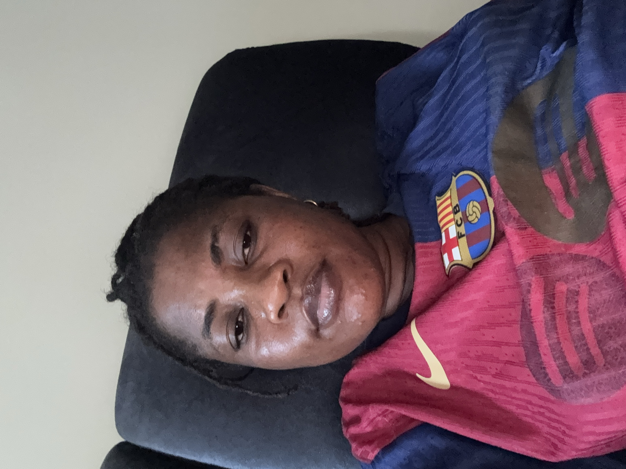 A medium shot captures a woman from the chest up, seated in a dark chair, against a plain, light-colored background. She has dark skin, dark hair styled in braids pulled back, and is wearing a sports jersey that is half red and half blue. The jersey features a Nike logo on the red side and a crest that reads "FCB" on the blue side. The woman's face is turned slightly towards the camera, and she has a neutral expression. Her lips are slightly parted and appear to be glossy. The overall lighting is bright and even. There are no discernible environmental features indicating a specific location in Ajara, Nigeria.