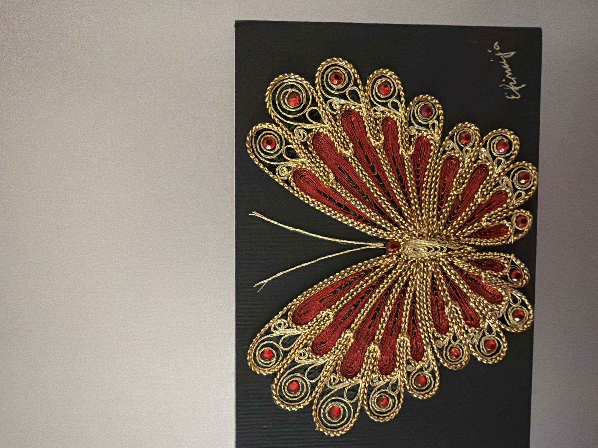A close-up, eye-level shot features a decorative butterfly artwork mounted on a black rectangular background. The butterfly is intricately crafted from red and gold threads, adorned with small, red, gem-like embellishments. The artwork is signed in script in the bottom right corner. The background is a textured black cardstock. The artwork is set against a plain, neutral-colored wall, with the top portion of the wall being a lighter shade and the bottom a darker shade. The lighting is even, with no distinct shadows. This artwork is located in Pazardzhik, Bulgaria.
