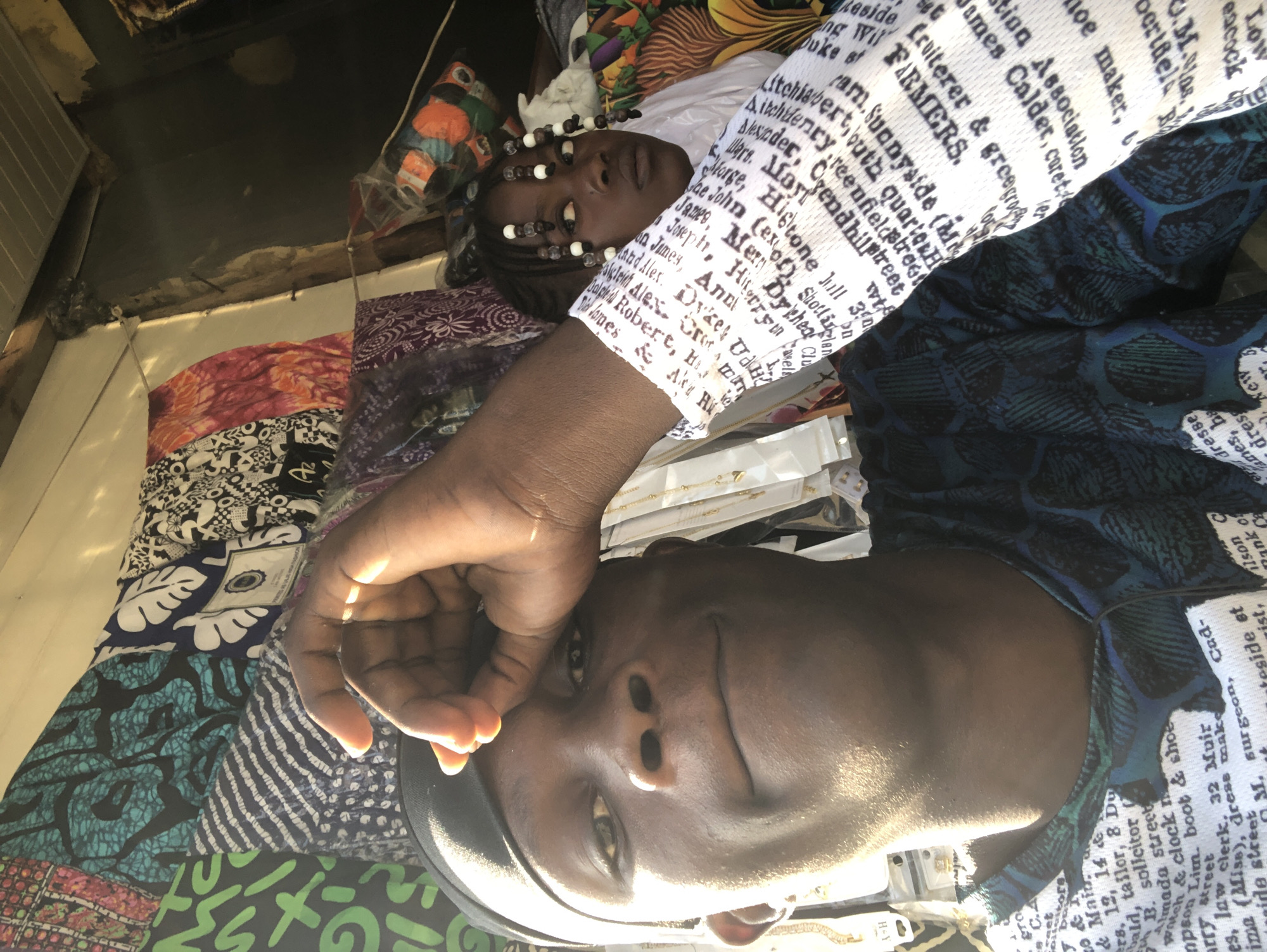 The image captures a close-up selfie taken by a young Black male in what appears to be a market stall in Ado, Nigeria. He is wearing a black durag and a dark blue t-shirt with a greenish-blue pattern. His right hand is raised, touching his forehead. To his right, a young Black girl is partially visible, with her hair styled in braids adorned with white beads. She is wearing a white long-sleeved garment with text printed on it, which is also visible on the male subject's arm. The background is filled with colorful fabrics and textiles, characteristic of a Nigerian market. The lighting suggests it is daytime, with sunlight casting shadows and highlights on the subjects and the environment. The overall impression is a candid moment captured within a vibrant, bustling marketplace.