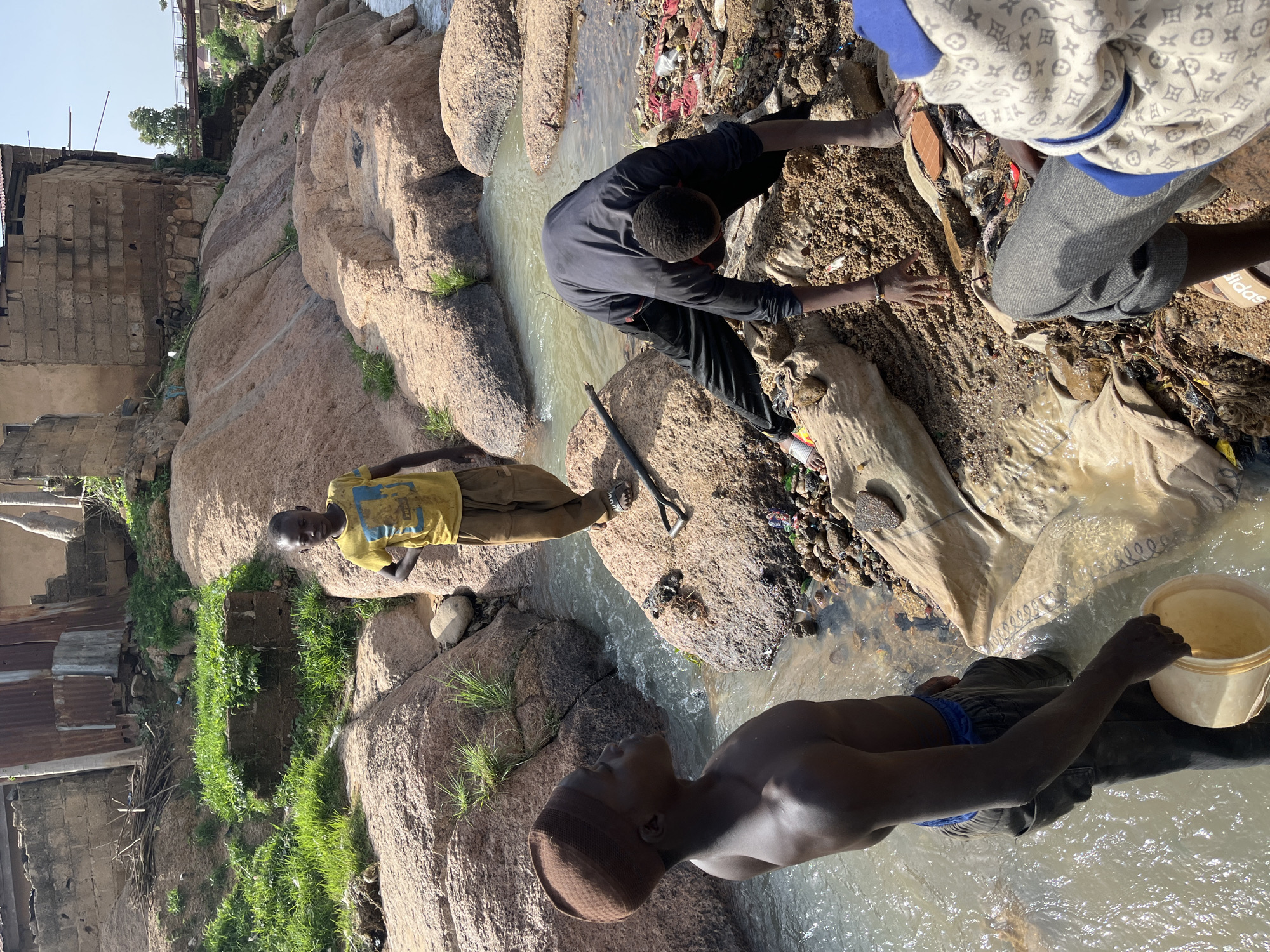 In Jos, Nigeria, at a rocky stream bed, four young Black boys are engaged in what appears to be a cleanup or resource extraction activity. One boy, wearing a yellow t-shirt and brown pants, stands on a rock, looking towards the camera with his hands on his hips. Another boy, bare-chested and wearing a brown kufi and dark pants, stands in the water, holding a pale yellow bucket. A third boy, dressed in a dark long-sleeved shirt and pants, is bent over, sifting through the rocky ground near the water's edge. The fourth boy, partially visible, is wearing a light-colored patterned hoodie and grey pants, and is seated with his hands near the ground. The environment is characterized by large, reddish-brown rocks, a shallow, flowing stream, and debris including plastic bags, fabric scraps, and other refuse scattered along the bank. In the background, there are structures made of concrete blocks and corrugated metal, suggesting a semi-urban or peri-urban setting. The overall scene depicts children working in a potentially polluted natural environment within the city.