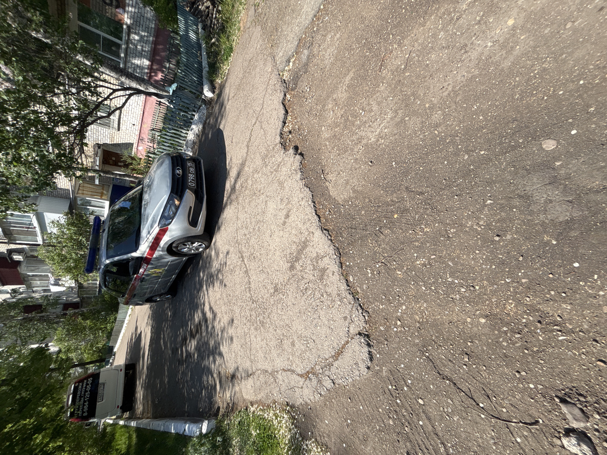 A street in Unknown, Unknown, is depicted in an oblique, close-up view. The roadway is made of asphalt and shows signs of wear and tear, with cracks and patches visible. A grey-colored Lada car, possibly a police vehicle due to the presence of a light bar, is parked on the left side of the frame. A section of a building is visible in the background, partially obscured by trees. A white van is positioned behind the car. Green foliage is present along the roadside.