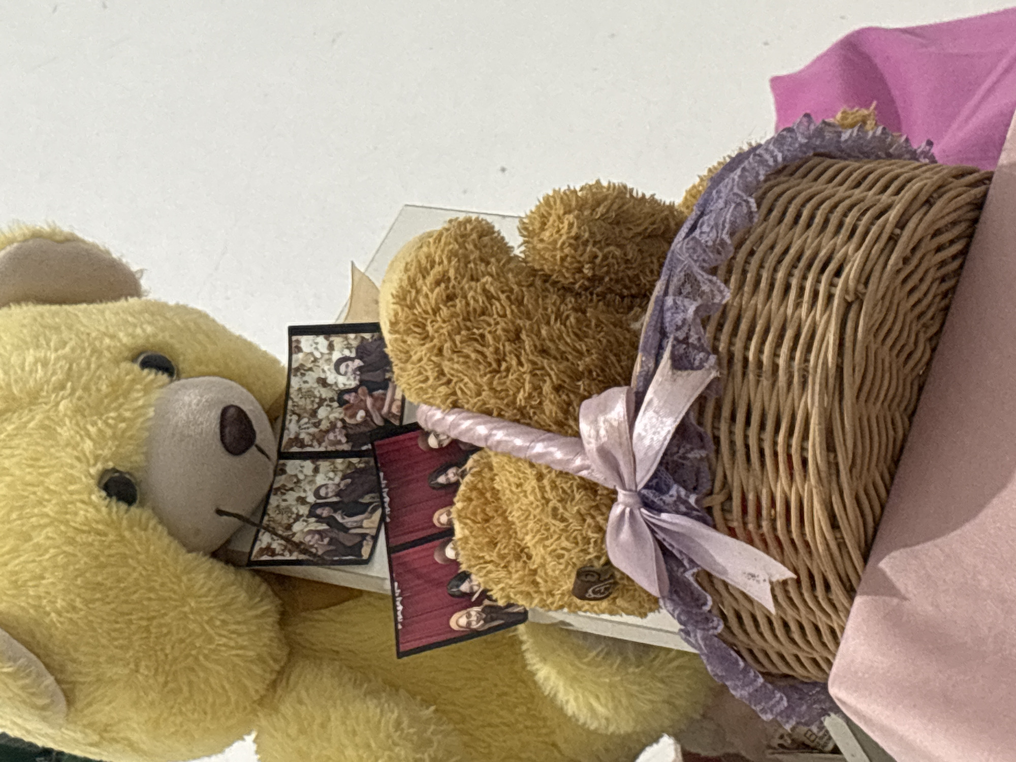 A close-up, slightly overhead shot captures two teddy bears and a woven basket. The larger, yellow bear is positioned behind and slightly to the left, its head visible from the forehead down to the snout. To the right of the yellow bear, and in front of it, is a smaller, brown teddy bear. This brown bear is situated in a woven basket that is decorated with a light purple lace trim and a light pink bow. The basket itself is filled with what appears to be red fabric or stuffing, and the brown bear is partially nestled within it. Several small, rectangular photographs are propped up behind the brown bear and in front of the yellow bear. These photos appear to show groups of people, with the one closest to the viewer showing five individuals standing against a red backdrop. The background of the image is a plain white wall. The scene is likely indoors, given the lighting and backdrop. There is no information available to determine the specific location within North Jakarta, Indonesia.
