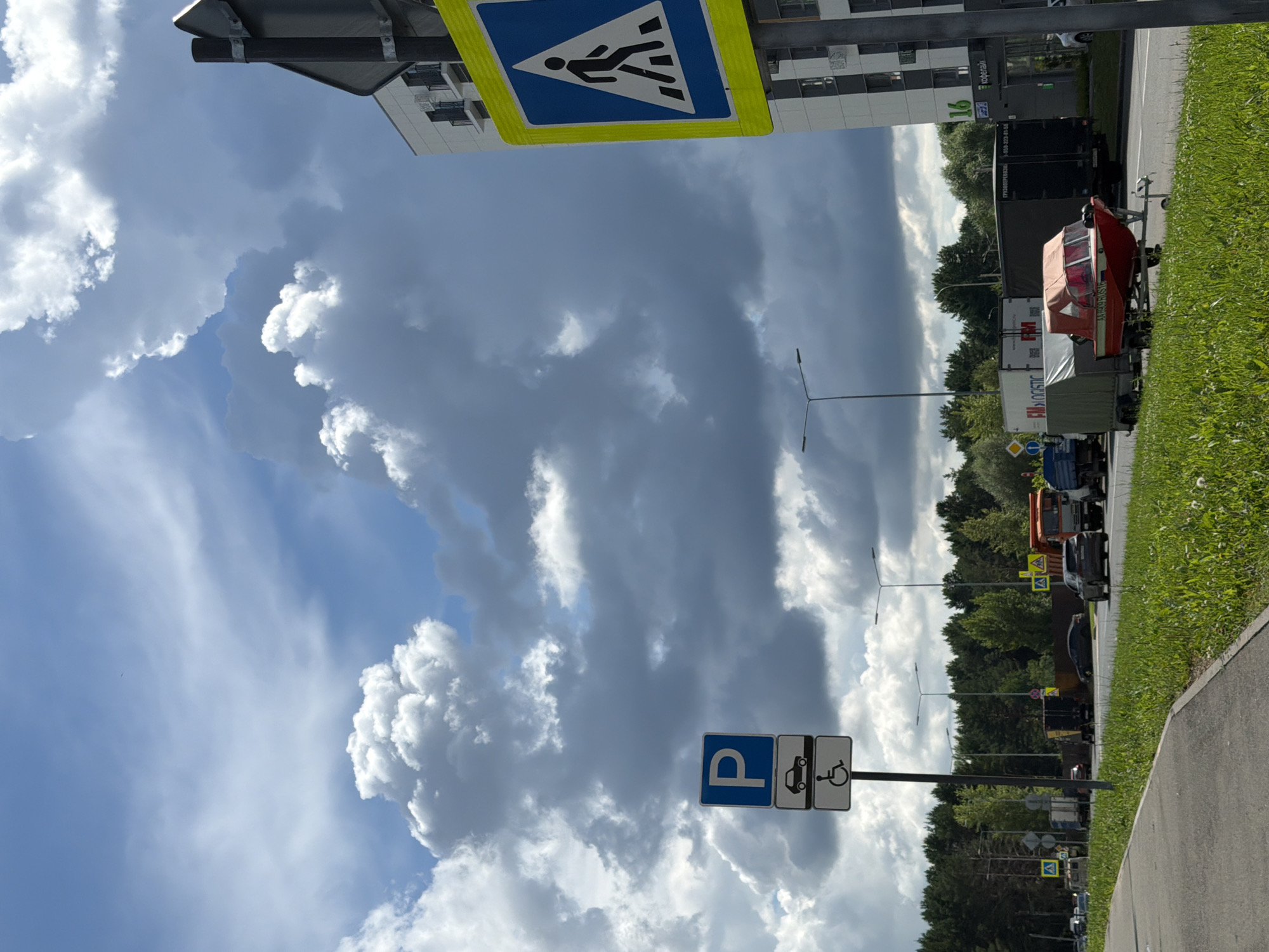 The image shows a street scene with a tall, modern apartment building on the right side, numbered "16" and bearing the sign "Кофеман" (Kofeman), indicating a coffee shop. In front of the building, a pedestrian crossing sign with a person walking is visible. To the left of the building, a tall pole displays two parking signs: one with a blue "P" for parking, and a lower sign showing a car and a wheelchair symbol, indicating disabled parking.
On the street, several vehicles are present. A red boat on a trailer is being towed by an unseen vehicle, positioned on the right side of the road. To its left are several trucks, including a large black truck and a white truck with "FM LOGISTIC" written on it. Further down the road, more trucks and cars are visible, suggesting a commercial or industrial area.
The road has a grassy median strip and a sidewalk on the left. The sky is dramatic with large, dark storm clouds mixed with patches of blue sky and bright white clouds. The environment appears to be a suburban or outskirt area of a city, given the presence of both residential buildings and commercial vehicles. The weather suggests an impending storm or recent rainfall. The image quality is clear, capturing details of the signs, vehicles, and the sky.