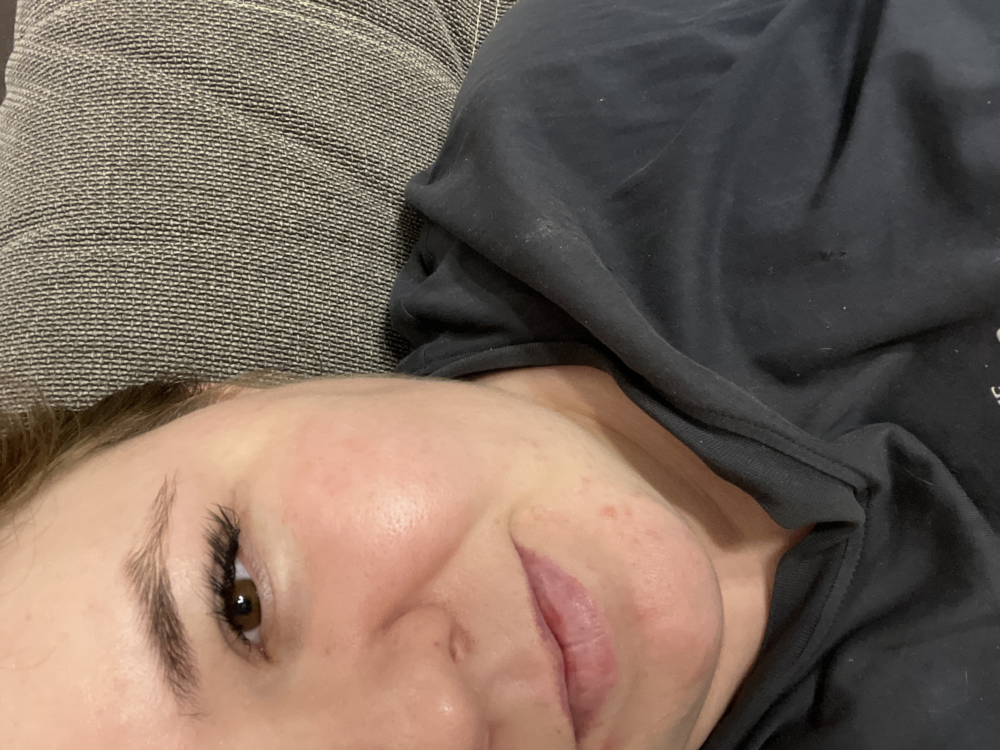 A close-up, angled shot shows a person lying down. The person has fair skin, a brown eye visible on the left, and is wearing a dark-colored shirt. The person appears to be on a grey fabric surface, possibly a couch or cushion. There is no information in the image to identify the location in Chaikovsky, Russia, or any actions or interactions.