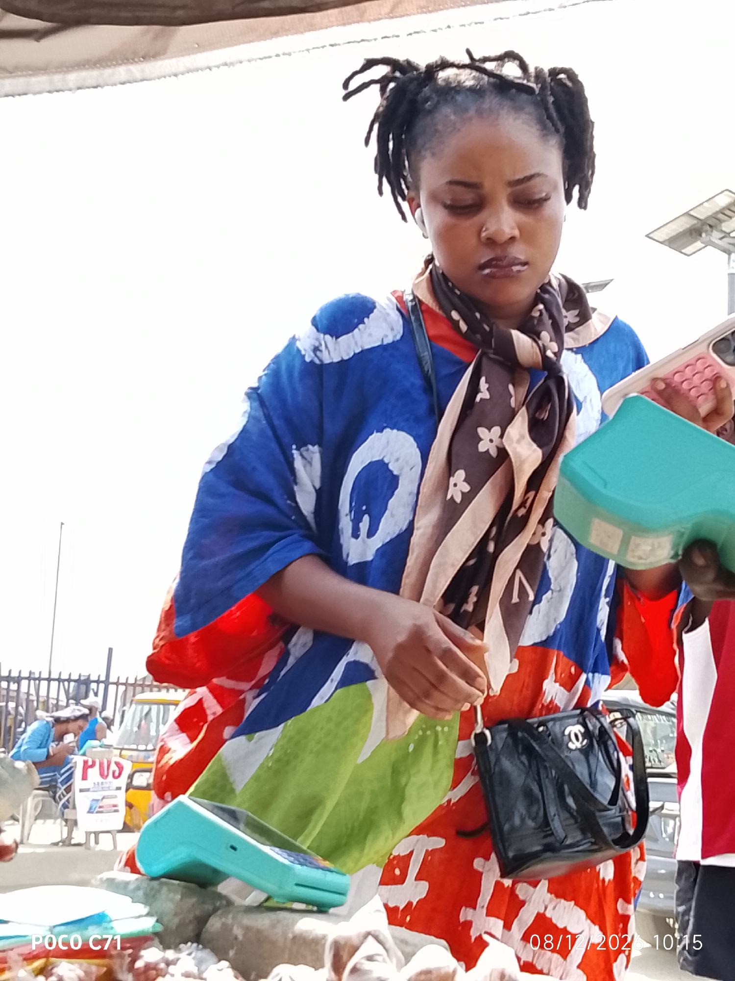 A woman with dreadlocks, wearing a colorful blue, red, and green patterned dress and a brown and black scarf, stands outdoors in Lekki, Nigeria. She is looking down at two mobile payment devices she is holding, one pink and one turquoise. A black Chanel purse hangs from her left shoulder. In the background, a yellow vehicle and a fence are visible. The image is timestamped "08/12/2020 10:15".