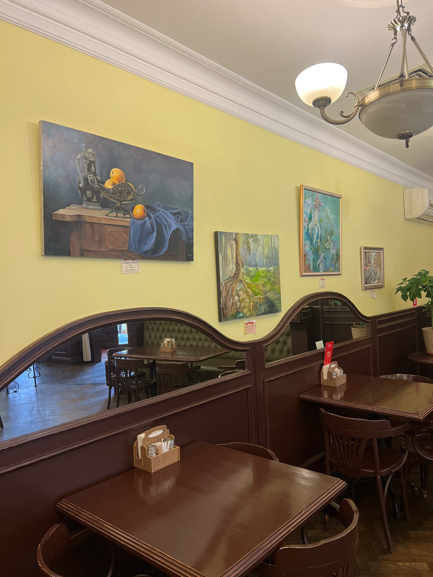 The image depicts the interior of a cafe or restaurant with a yellow painted wall and decorative white crown molding. Several paintings are displayed on the wall. One painting on the left is a still life of a silver pitcher and fruit on a wooden surface, draped with blue fabric. To its right, a painting shows a forest scene with large tree roots. Further right is a vertical painting of irises. On the far right, a smaller framed painting depicts a street scene. Below the paintings, dark brown wooden booth seating with tufted upholstery is visible, separated by a mirrored partition. In the foreground, dark wooden tables and chairs are arranged within the booths. A wooden condiment caddy with napkins and packets is placed on one table. A chandelier hangs from the ceiling, providing light. The cafe appears to be empty of people. The location is identified as "Dux" in Kaliningrad, Russia, as indicated by a sign on one of the tables in the reflection.