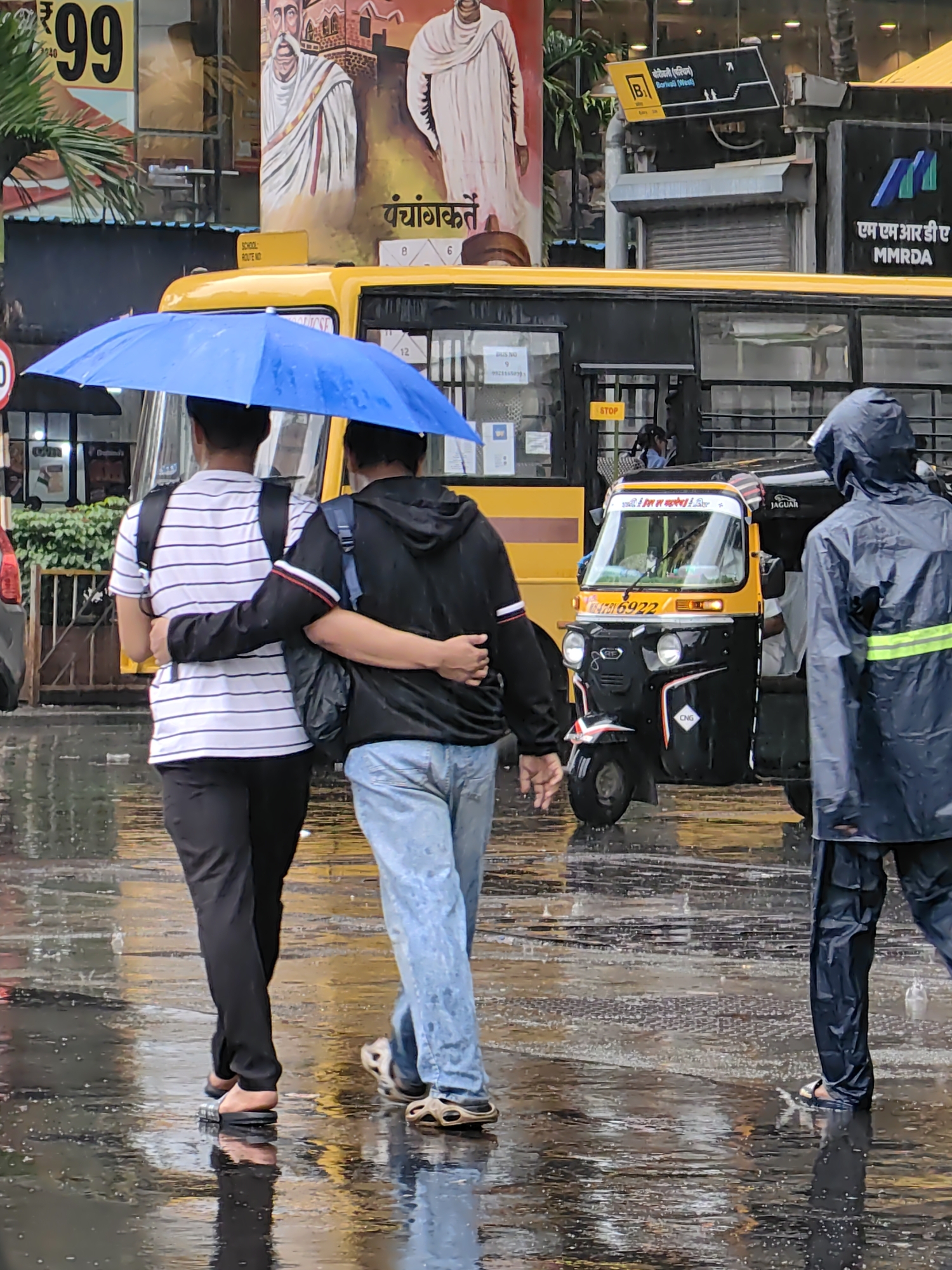 Two individuals, one in a black hoodie and the other in a striped t-shirt, walk under a blue umbrella on a wet street in Mumbai, India. The person in the black hoodie has their arm around the other person's shoulder. A third person, wearing a dark blue rain jacket, walks away from the camera. In the background, a yellow school bus and a black and yellow auto-rickshaw are visible. The street is reflecting the surrounding lights and vehicles, indicating it is raining. The scene suggests a typical monsoon day in Mumbai.