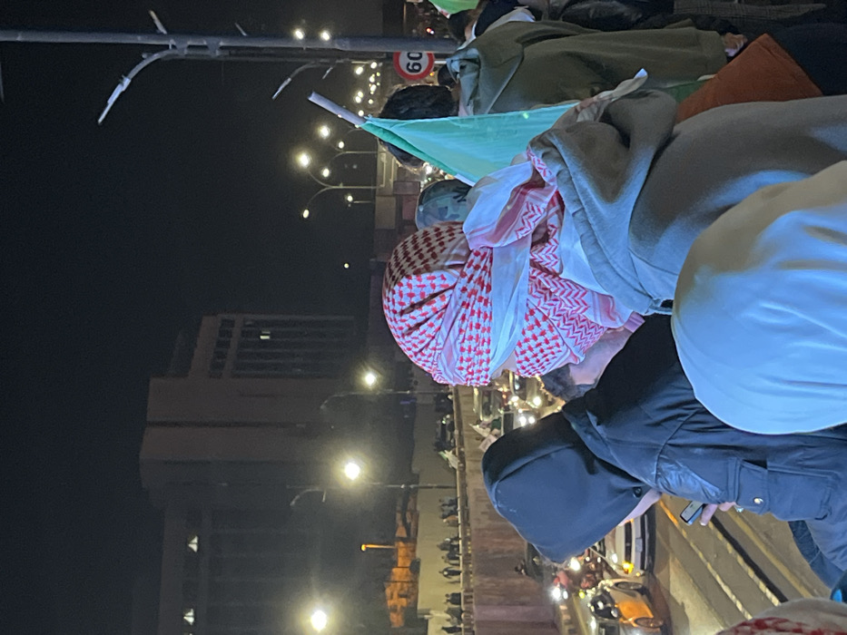 A nighttime gathering of people is visible. Some individuals are draped in traditional red and white patterned cloths. A green flag on a pole is visible. The background includes buildings and streetlights. There are several people wearing hooded jackets.