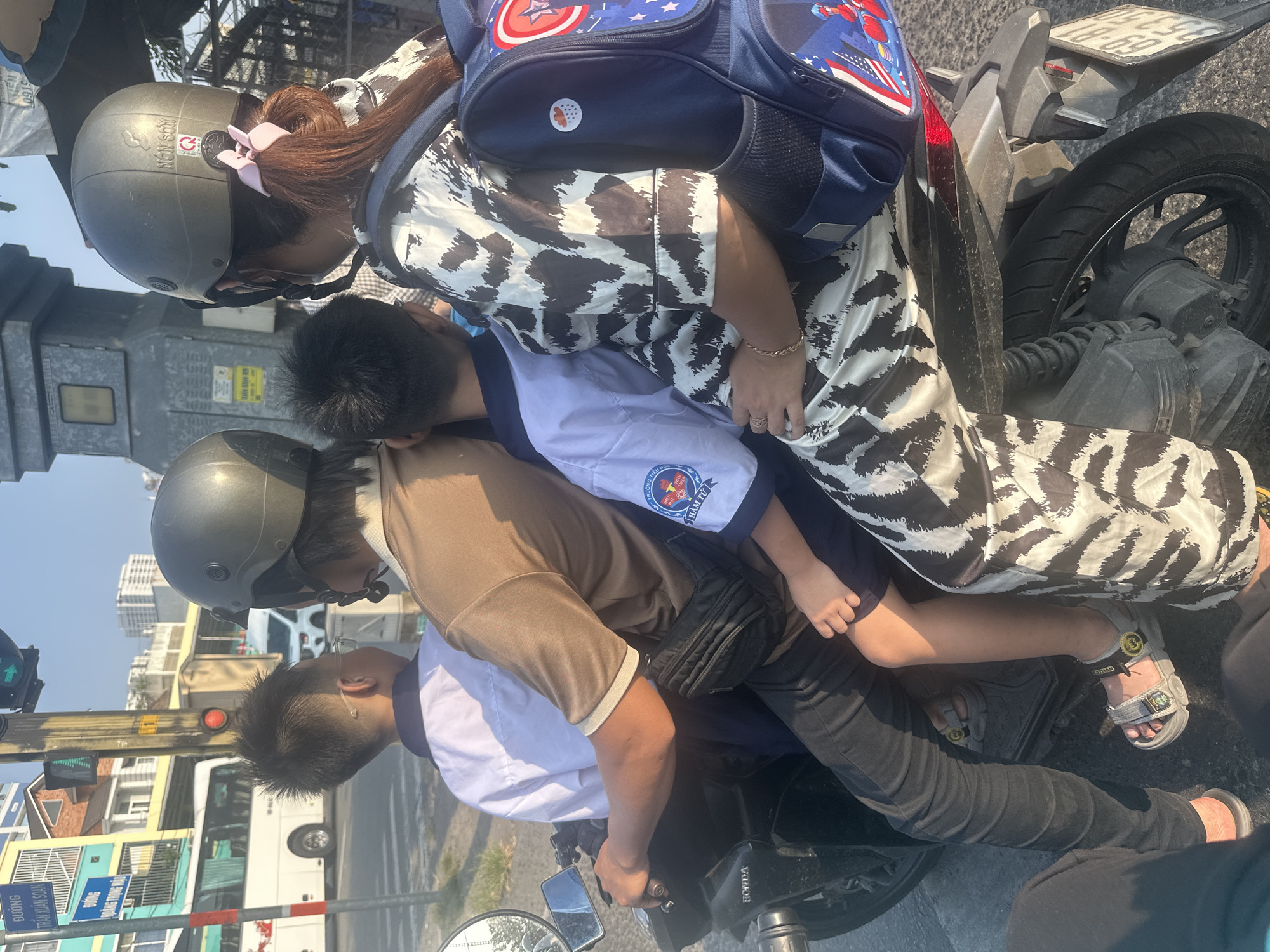 A motorcycle is carrying three individuals on a city street in Ho Chi Minh City, Vietnam. The driver is an adult wearing a brown shirt, black jeans, black sandals, and a gray helmet. Two children are riding behind the driver, with the younger child sandwiched between the driver and an adult wearing a white and brown patterned shirt and a gray helmet. The younger child has on sandals and a school uniform. The adult in the back is wearing a backpack with a Captain America design. The motorcycle, identified as a Honda, is situated near a traffic light. Buildings are visible in the background. The license plate of the motorbike reads 59-L8 181.69. Street signs are also visible, including one that reads "DUONG TRAN XUAN SOAN." Another sign reads "TRUONG TIEU HOC HAM TU," and contains the symbol of the school and its name.