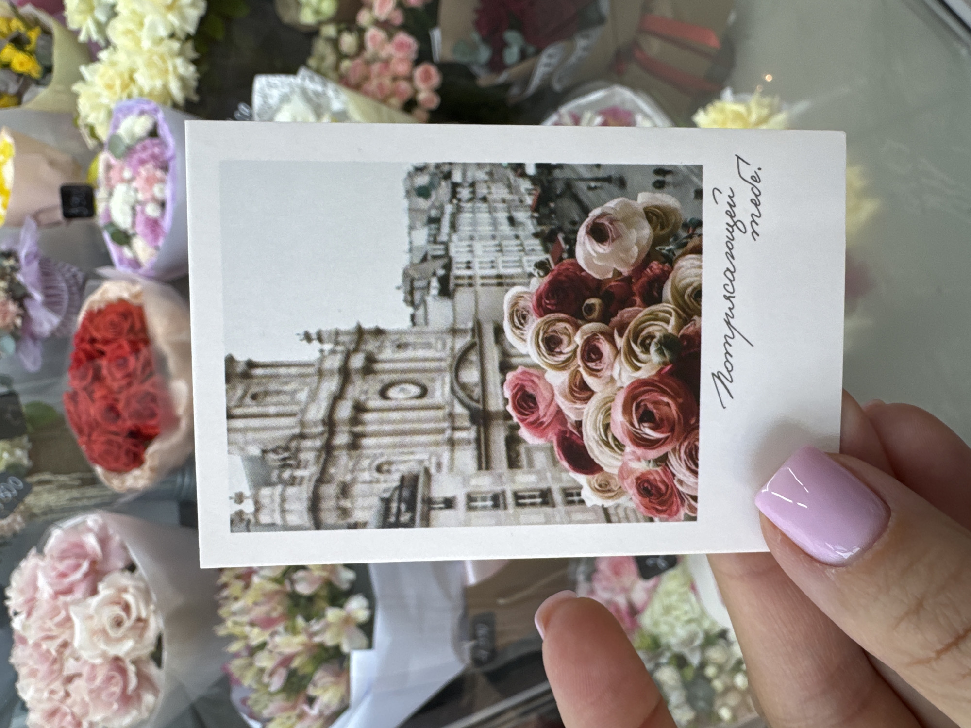 A medium shot captures a hand holding a small, rectangular photograph against a backdrop of various flower bouquets. The hand, with light skin and a light purple, glossy nail polish, subtly obscures the bottom portion of the card. The photograph within a photograph is a grayscale image of a building with intricate architectural details, juxtaposed with a vibrant bouquet of roses in varying shades of red, pink, and cream. Handwritten text in a cursive script is visible along the right edge of the card, partially covered by the hand. Surrounding the hand and the card are multiple flower bouquets, adding depth and color to the composition. No location is discernible.