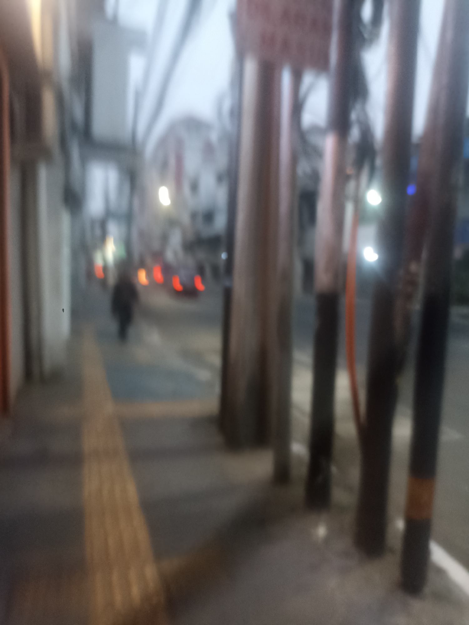 The image depicts a blurry outdoor scene, likely a street in Bandung City, Indonesia. In the foreground, several thick, vertical poles are visible, some with orange bands. To the left, a building facade is partially visible, with a sidewalk running alongside it. Further down the street, a lone figure, appearing to be a pedestrian, is walking away from the camera. The street is lined with what seem to be streetlights or illuminated signs, and there are blurred shapes of vehicles with visible red taillights in the distance, suggesting evening or night time. The overall impression is one of a street scene with ambient lighting and limited visibility due to blurriness.