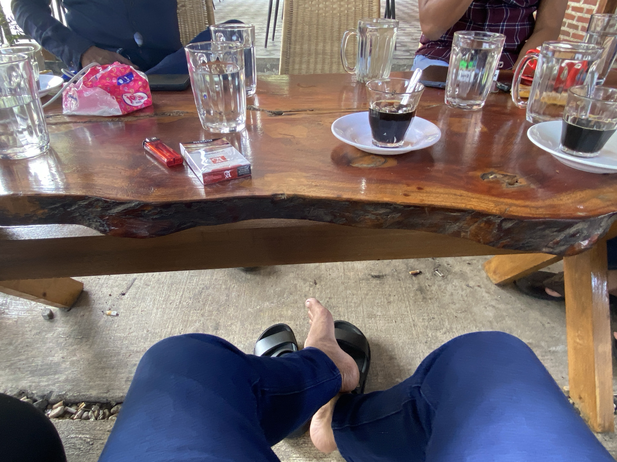 This image depicts a scene at a table, likely outdoors, in Aluepiet, Indonesia. The focus is on a wooden table with various items on it and the lower body of a person wearing blue pants and black sandals.
On the table, there are several clear glasses, some filled with water, others with dark liquid appearing to be coffee. A small white saucer holds a glass of coffee with a spoon. A pack of cigarettes and a red lighter are also present. A pink and white patterned wrapper, possibly for tissues or snacks, is near the glasses. In the background, parts of people can be seen, including a person wearing a dark shirt and another wearing a maroon and white checkered shirt. The table appears to be set up in a casual outdoor area, possibly a cafe or roadside eatery, with concrete flooring visible below. There are also some discarded cigarette butts on the concrete. The lighting suggests it is daytime.