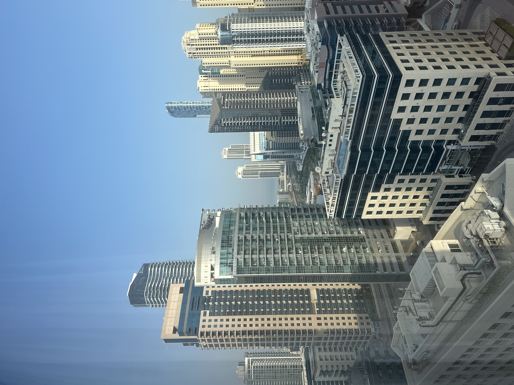 The image presents an aerial view of a modern cityscape, specifically capturing the Dubai Marina area in Dubai, United Arab Emirates. The scene is dominated by a dense collection of high-rise buildings, characterized by contemporary architectural designs and varying facade materials, including glass, concrete, and metallic finishes.
The buildings exhibit a range of heights and forms, with many featuring multiple balconies and windows, indicating residential or commercial use. Some structures have unique geometric shapes and curved exteriors, highlighting the advanced construction and design prevalent in the area. Several prominent buildings are identifiable by their signage, such as "EMAAR" and "SPARKLE TOWERS," which are well-known entities in Dubai's real estate and development sector.
In the foreground, a few buildings are closer to the viewer, offering a detailed look at their rooftops. These rooftops are equipped with various mechanical systems, including air conditioning units, ventilation systems, and satellite dishes, suggesting the functional infrastructure supporting these structures. One rooftop features a swimming pool, indicating amenities provided for residents or occupants.
The background reveals a glimpse of the sea or a large body of water, with a bridge or elevated roadway connecting different parts of the city. The sky is a clear, bright blue, indicative of fair weather, and the overall lighting suggests it is either morning or late afternoon, given the soft shadows cast by the buildings.
No people are visible in the image, and there are no specific actions or interactions to describe. The focus is solely on the urban landscape and its architectural components. The scene is a testament to the rapid development and impressive skyline of Dubai, particularly in the Marina district, known for its concentration of luxury residential and hospitality towers.