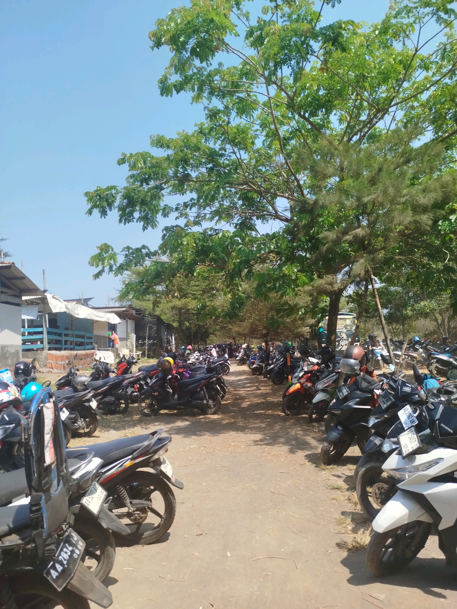 The image depicts a crowded parking area for motorcycles in Kenoyajaya, Indonesia, likely near a beach or recreational area. The scene is dominated by a large number of parked scooters and motorcycles, filling a dirt and gravel lot. In the background, a row of simple structures, possibly food stalls or small shops, are visible, with some featuring open sides and covered roofs. Lush green trees with dense foliage provide shade over parts of the parking area and extend towards the bright blue sky. Some people are present, but they are not the primary focus and are largely obscured or indistinguishable among the parked vehicles. The overall impression is one of a bustling, outdoor commercial or recreational space.