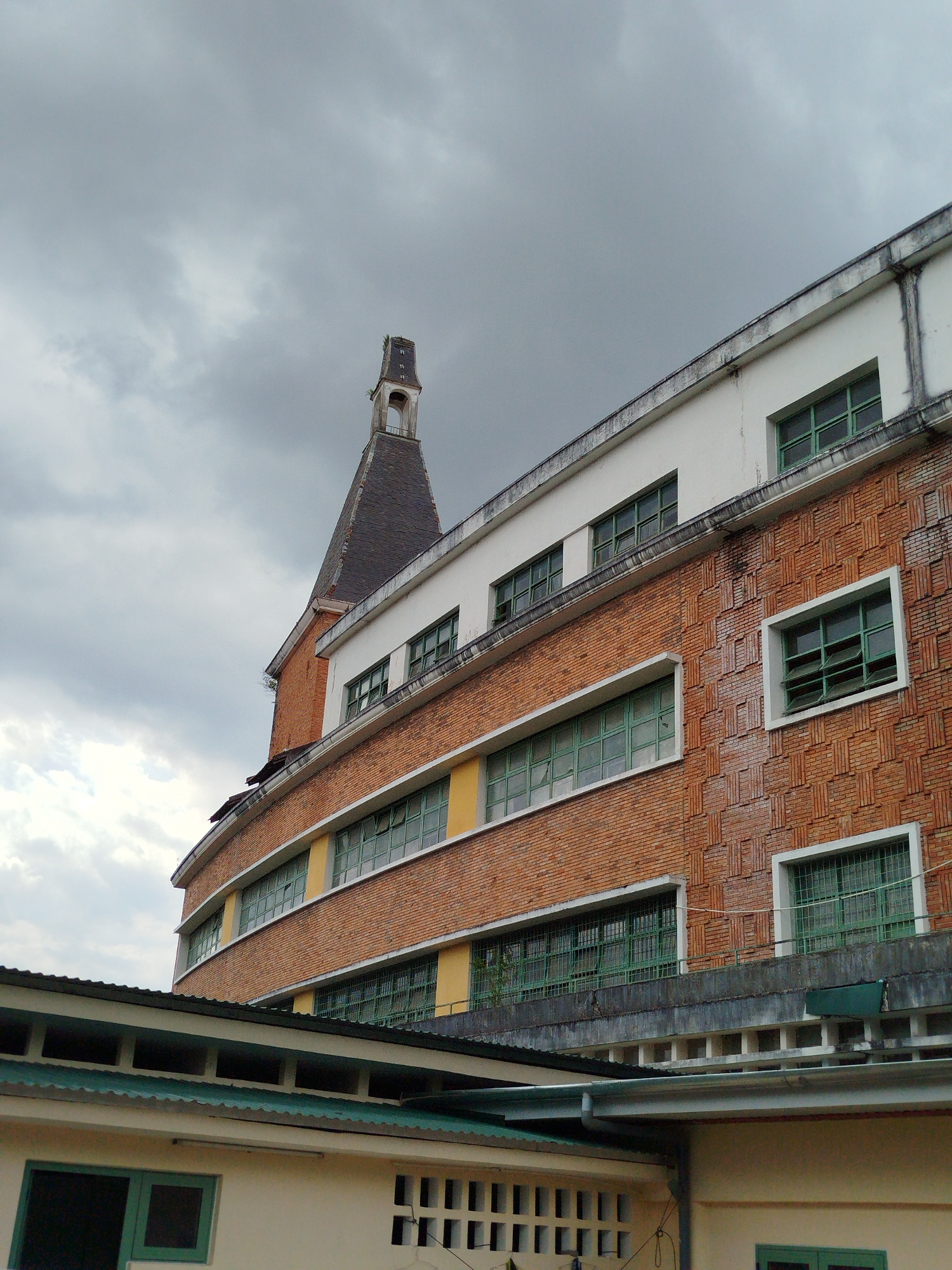 The image shows a low-angle view of the Lycée Yersin Da Lat building in Da Lat, Vietnam. The building features a combination of red brick and white sections, with rows of green-framed windows. The brickwork has a textured pattern. A tall, narrow spire-like structure rises from the building's roof. The sky is overcast with dark gray clouds. In the foreground, there's a lower roof section with a green corrugated metal covering, and the exterior wall of another building with green window frames and decorative vertical slits.