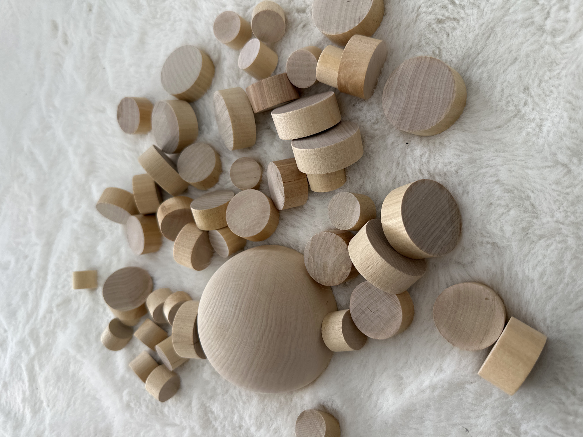 The image shows an arrangement of small, unfinished wooden cylinders and one large wooden disc on a white, fluffy surface. There are no people or other objects visible. The environment is undefined beyond the white surface, making location identification impossible.