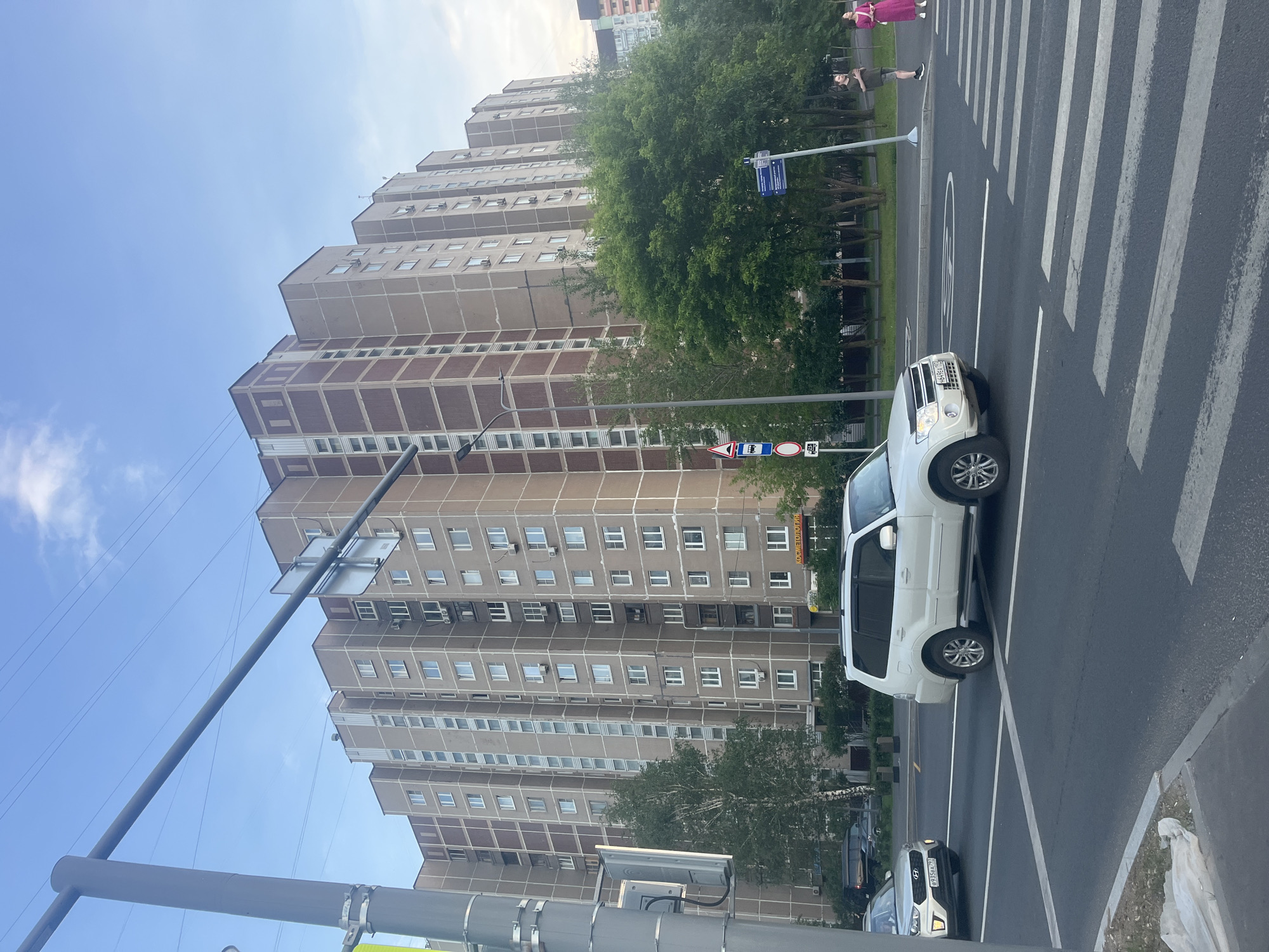 This is a street-level, medium-shot photograph taken in Moscow, Russia, on a clear day with a bright blue sky and scattered clouds. The scene is dominated by a large, multi-story apartment building with a light brown facade and darker brown trim around the windows. The building appears to be a typical Soviet-era construct, featuring many identical windows and balconies. In the foreground, a white SUV is stopped at a pedestrian crossing on a paved road. Another car, a silver sedan, is approaching the crossing from the left. A pedestrian crossing sign and a bus stop sign are visible on the sidewalk to the left of the road. Trees line the sidewalk in front of the apartment building, and a few individuals are visible on the sidewalk in the distance, near a street sign. The overall impression is a typical urban street scene in Russia.