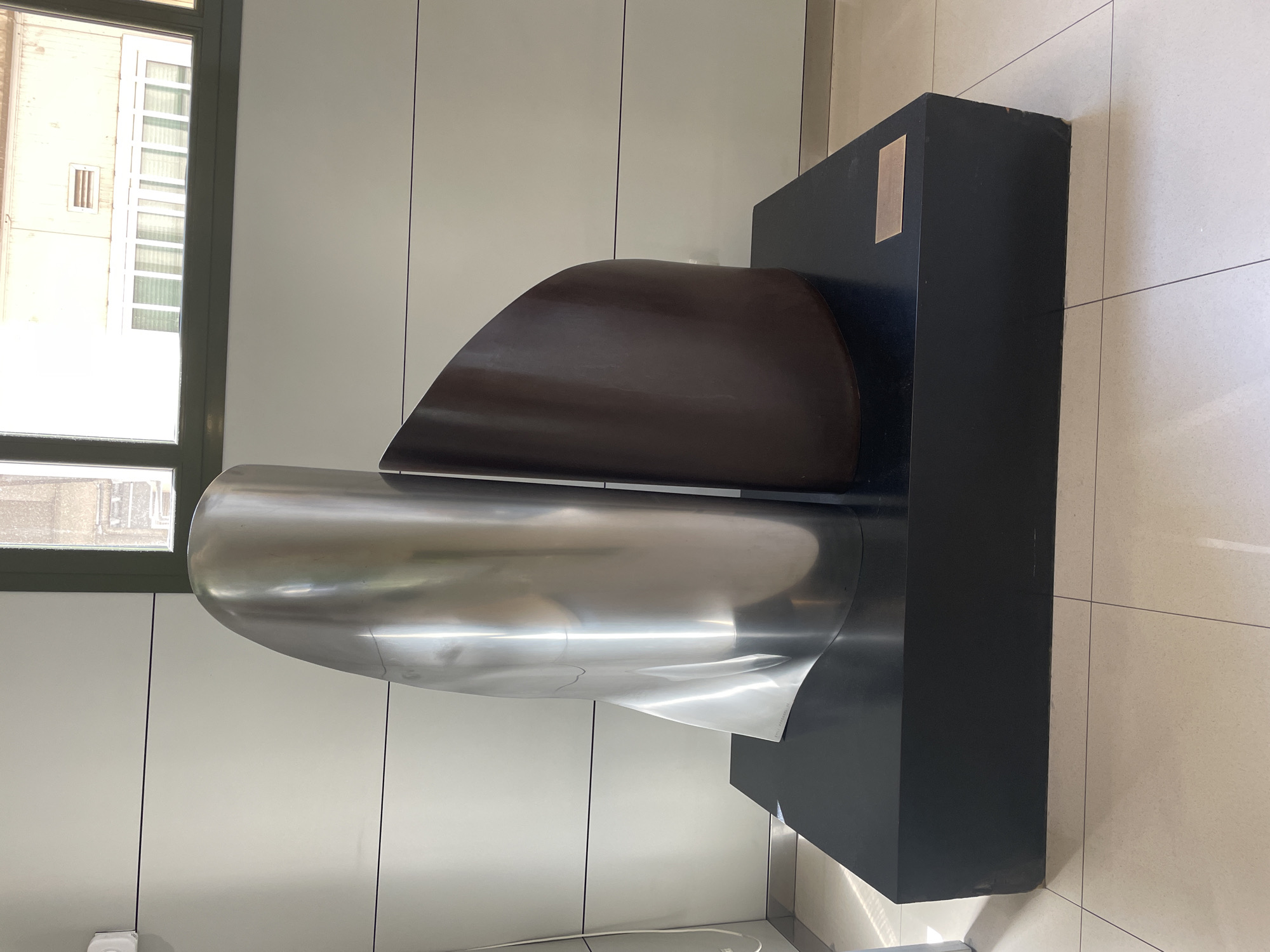 A contemporary sculpture composed of two large, abstract, curved forms stands on a black rectangular pedestal. The form on the left is polished silver, reflecting light and its surroundings. The form on the right is a dark, reddish-brown, with a matte finish. A small, rectangular plaque is affixed to the front right side of the pedestal. The sculpture is placed in an indoor environment with pale walls and a tiled floor. A window with green framing is visible in the upper right portion of the image, showing buildings and sky outside. The scene appears to be a public space, possibly a museum or a gallery, within Barcelona, Spain.