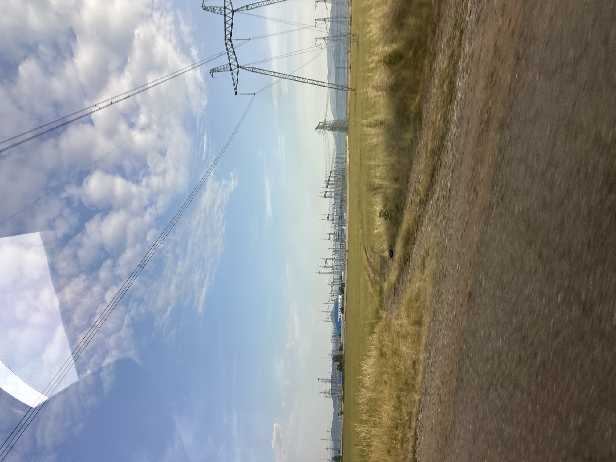The image displays a landscape with a clear sky filled with scattered clouds. Tall power lines and pylons stretch across the frame, indicating an area with significant electrical infrastructure. The foreground on the right shows a dirt or gravel road, with dry grass and vegetation along its edge, suggesting a rural or undeveloped setting. In the distance, more power lines and structures are visible, along with rolling hills or mountains under a hazy sky. The overall scene conveys a sense of open space and industrial presence within a natural environment. The lighting suggests it is daytime, likely late morning or early afternoon given the bright sky. There is no visible text in the image.