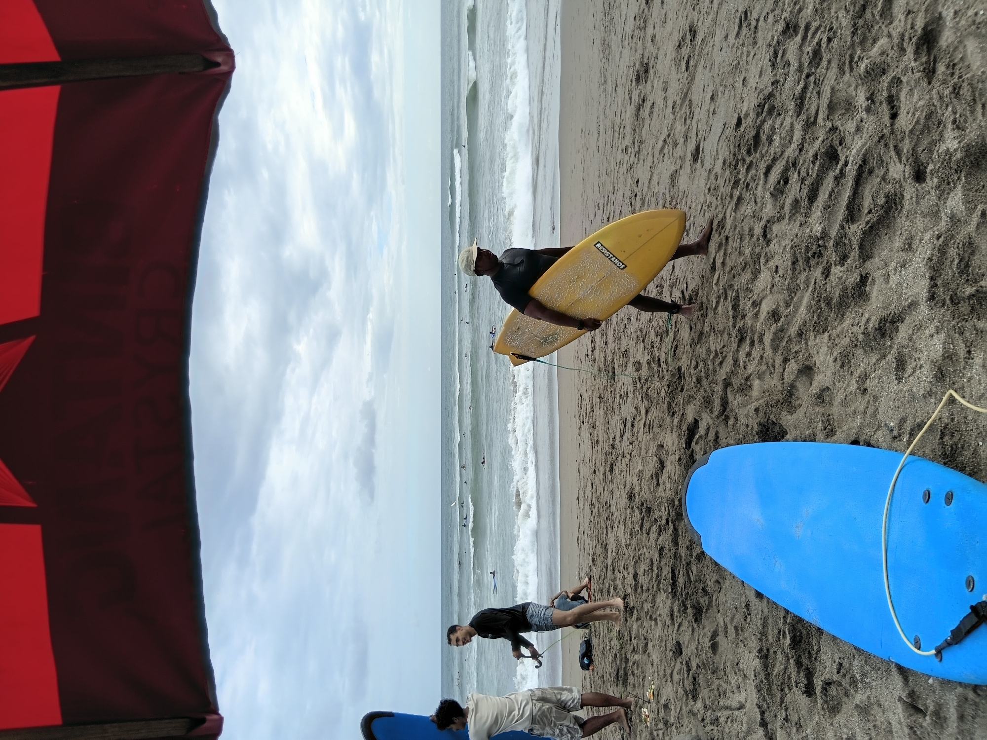 In North Kuta, Indonesia, a group of surfers are gathered on a sandy beach under a cloudy sky. In the foreground, a bright blue surfboard lies flat on the sand. To the left, a young man wearing a light-colored t-shirt and patterned shorts stands holding a blue surfboard. Next to him, another young man in a black long-sleeved shirt and patterned shorts is talking while holding a leash. Further back on the beach, a man wearing a dark t-shirt and a light-colored hat carries a yellow surfboard. In the background, the ocean waves are breaking, and several surfers are visible in the water. The overall scene depicts a typical day at a surfing beach in Bali, Indonesia.