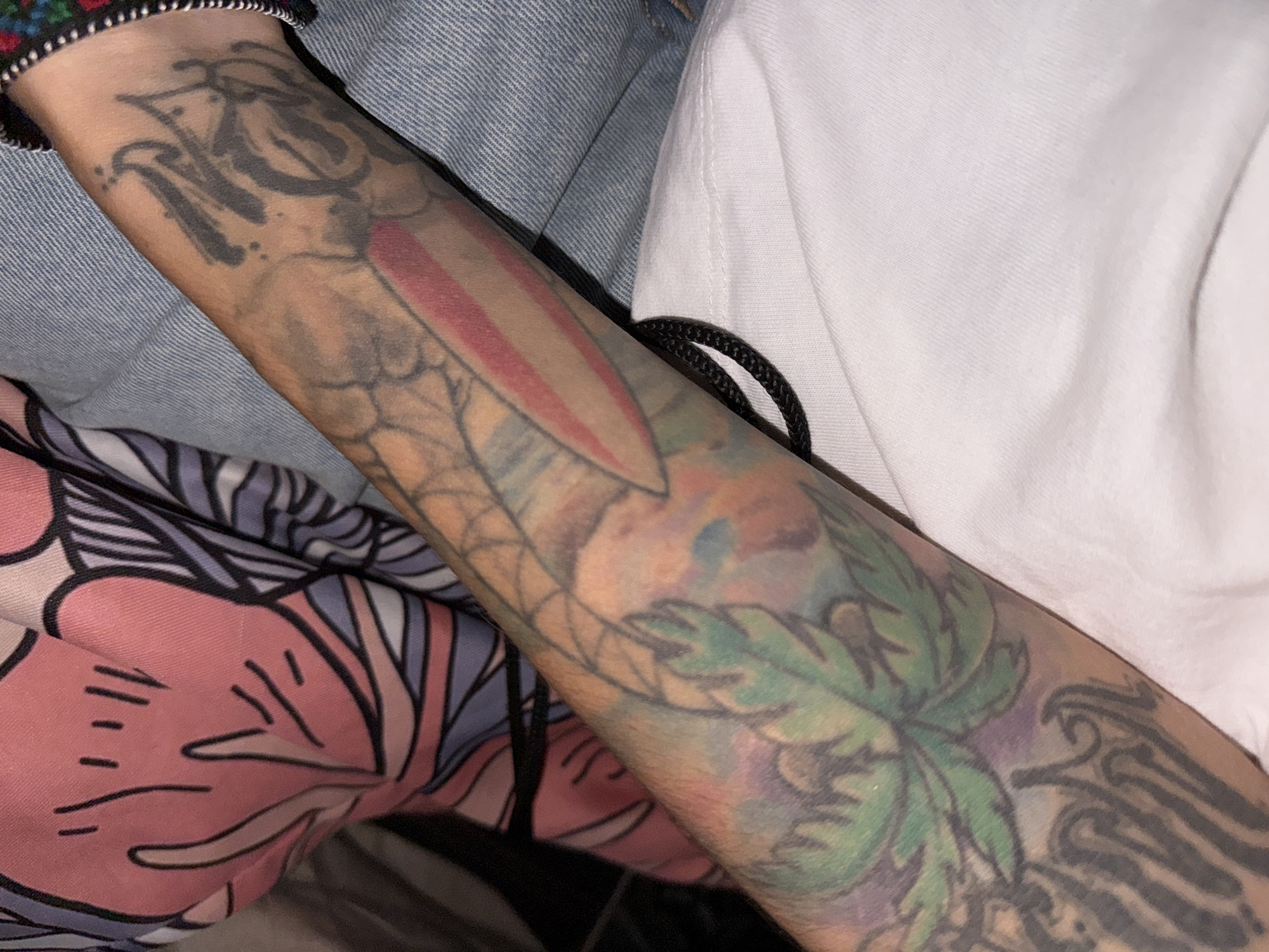 A close-up shot features a person's tattooed forearm, angled diagonally across the frame. The arm is extensively covered with multiple tattoo designs. On the upper part of the forearm, near the wrist, are dark grey, script-like letters that are difficult to definitively decipher, possibly forming "NO" or "NCS." Below these letters, a distinctive red and white striped element, resembling a knife blade or a stylized flag, is visible, surrounded by a faint, web-like or scaled pattern. Further down the arm, a prominent green leafy design with watercolor-like splashes of purple and blue hues is present. Towards the lower portion of the forearm, darker, more abstract tribal-style patterns or wings can be seen.
The arm is set against a backdrop of various fabrics: a textured blue denim material (likely a jacket) is visible on the upper left, and a plain white t-shirt or garment occupies the right side of the frame. A black braided cord is nestled between the arm and the white fabric. In the lower left, a soft pink and purple fabric with a stylized floral or abstract pattern is visible, possibly part of another piece of clothing or a blanket. The image is a close-up, offering no clear indication of a specific activity or the time of day.