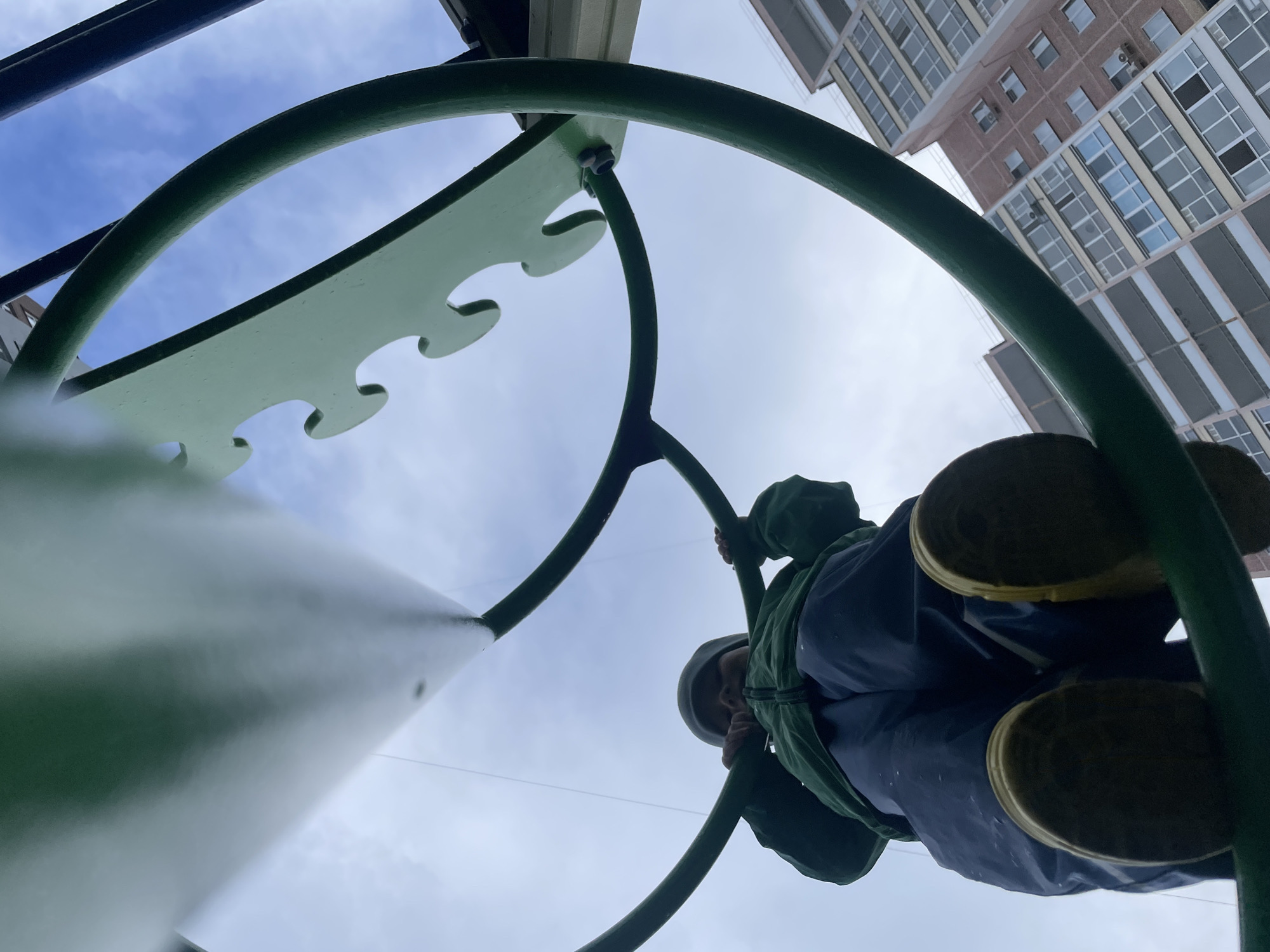 An extreme low-angle shot captures a person playing on green playground equipment against a bright, cloudy sky and a distant tall building. The perspective is from directly beneath the structure, looking upwards.
The main subject is a person, likely a child, seen from the waist down and partially from the head/hood, appearing to be either climbing or sliding. They are dressed in a dark green hooded jacket, dark blue pants or overalls, and dark footwear with light-colored soles. Their face is obscured, but their hands are visible grasping the green metal bars of the playground.
The dominant objects in the foreground are the dark green, curved metal bars and lighter green, flat panels with wavy edges, which form part of the playground equipment. A broad, light green flat surface, possibly a slide or ramp, extends from the lower left foreground into the image.
In the upper right background, a multi-story building with a reddish-brown brick facade and numerous windows is visible, indicating an urban or suburban setting. The sky is bright with white clouds, and patches of blue are visible. The time of day appears to be daytime, possibly overcast or partly cloudy. There is no visible text in the image.