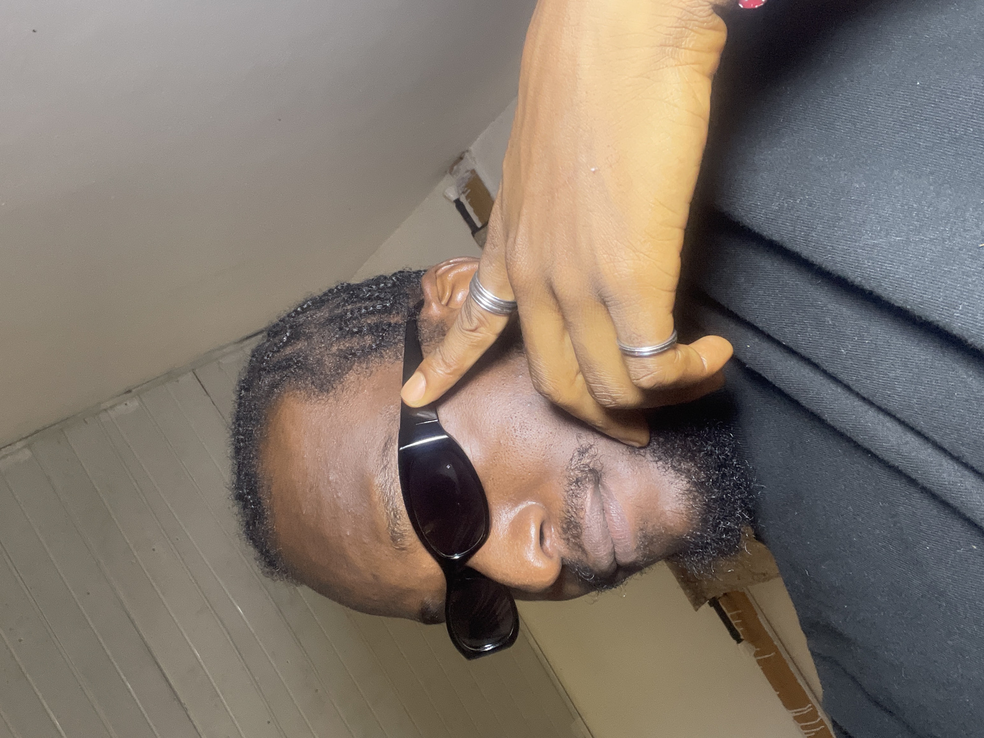 The image shows a close-up, low-angle shot of a Black man wearing sunglasses and a black garment. He has braids and a beard, and is wearing two silver rings on his right hand. His hand is positioned near his face, obscuring part of it. The background appears to be an indoor setting with cream-colored walls and wooden paneling. The lighting suggests it might be daytime, but the interior setting makes it difficult to determine the time of day. There are no obvious location cues to suggest it is specifically Ban, Nigeria, beyond the provided context. The man's expression is not clearly visible due to the sunglasses and hand. The overall mood is somewhat enigmatic.