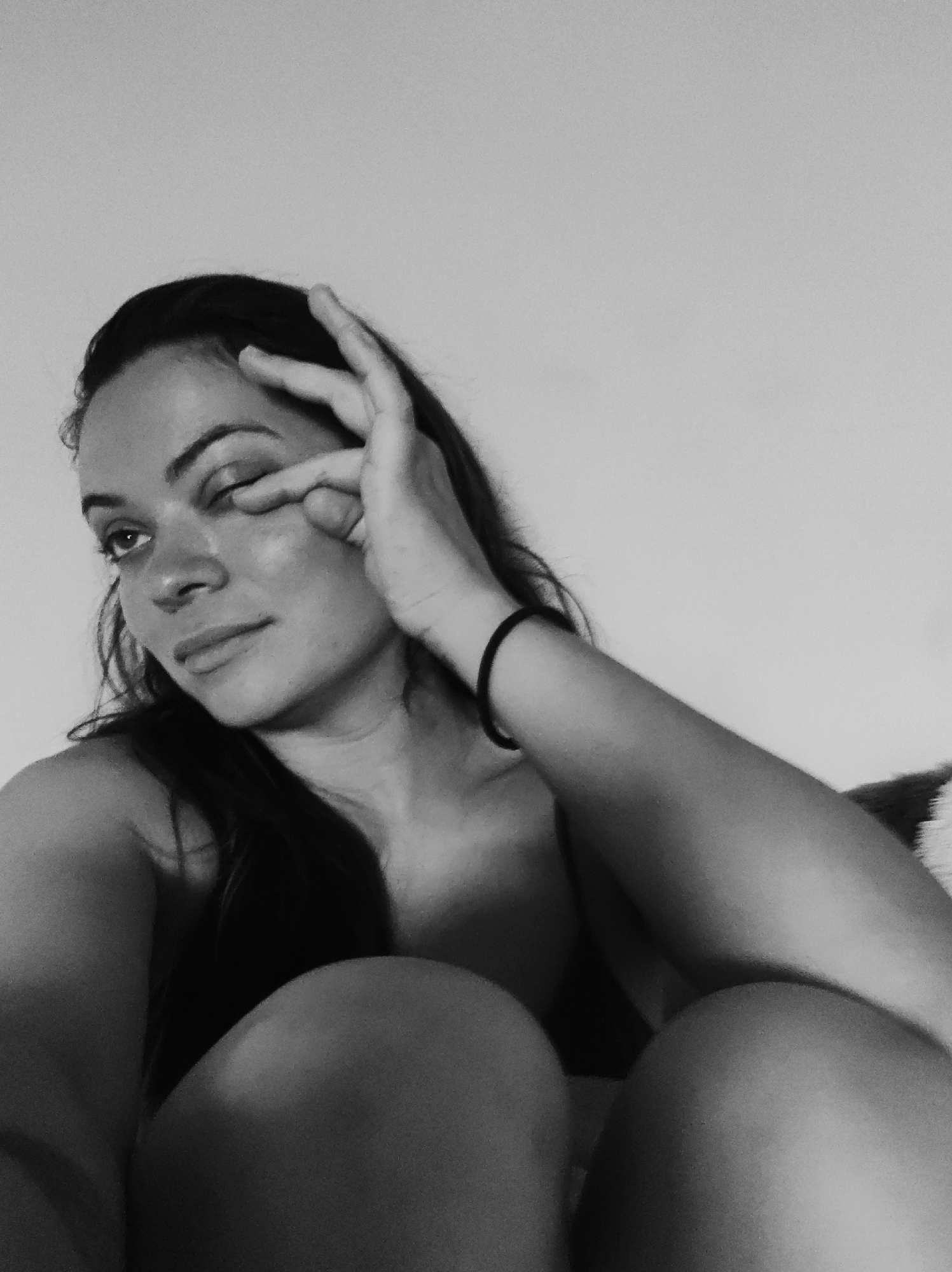 This black and white photograph captures a young adult female from a low-angle perspective, seated or reclined indoors in the city of Unknown, Unknown. The subject, with long dark hair, is looking upwards and to her left with a soft, contemplative expression; her lips are slightly parted. Her left hand is raised to her face, with the index and middle fingers extended towards the outer corner of her left eye, while the other fingers are curled. A black elastic hair tie is present on her right wrist. Her shoulders and the upper part of her chest are visible, suggesting minimal upper body clothing. Her knees are drawn up, occupying the lower portion of the frame. The background is a plain, light-colored wall, providing no additional environmental details.