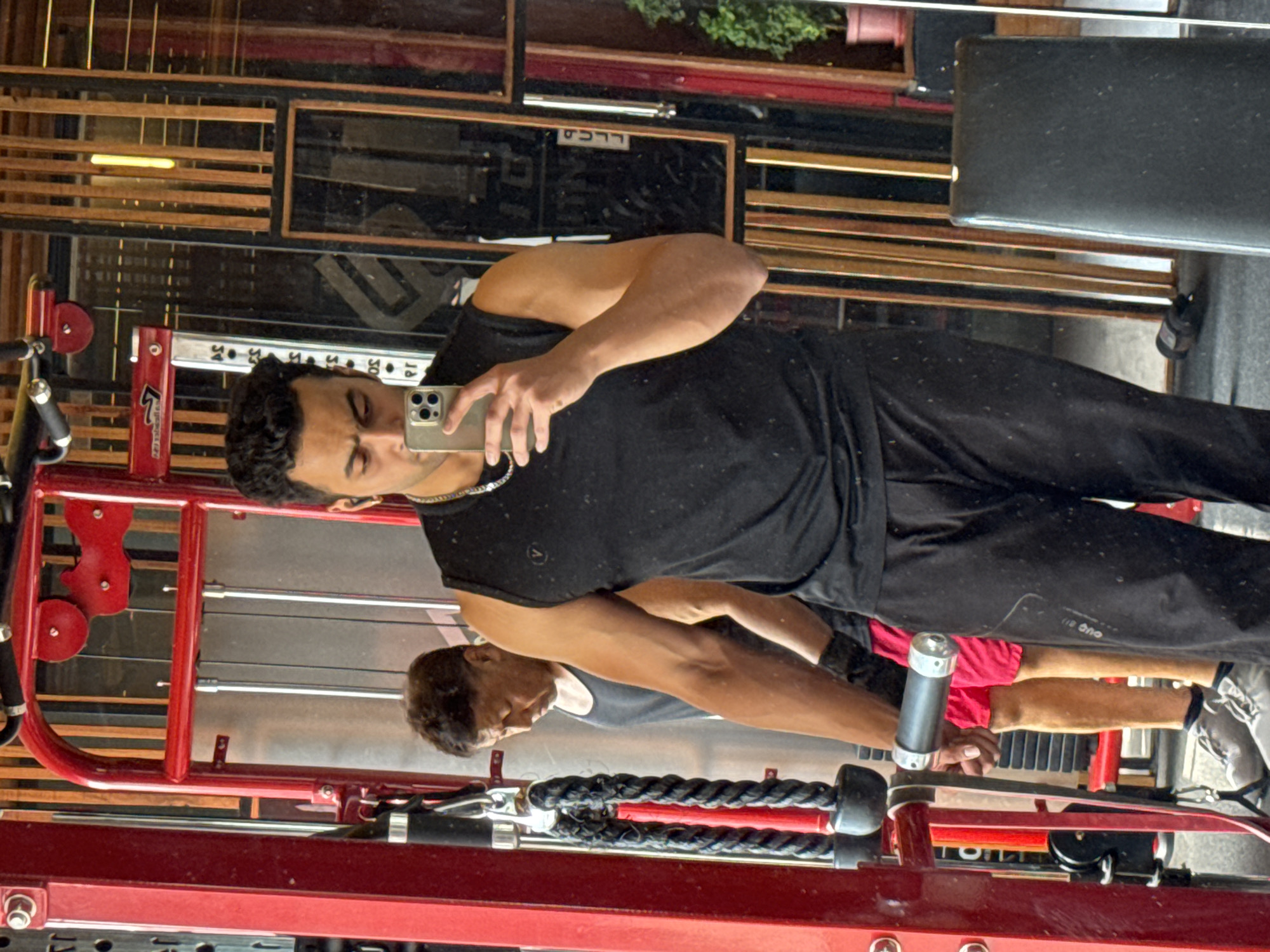 A man in a black sleeveless shirt and black track pants takes a mirror selfie in a gym. He is holding a smartphone in his right hand. In the reflection of the mirror, another man in a black tank top and red shorts is visible in the background, standing near a weight machine. The gym appears to have red and metal gym equipment. The background also shows wooden panels and other reflections. The scene suggests a modern gym environment, likely in a city like Kolkata, India, given the visual cues.