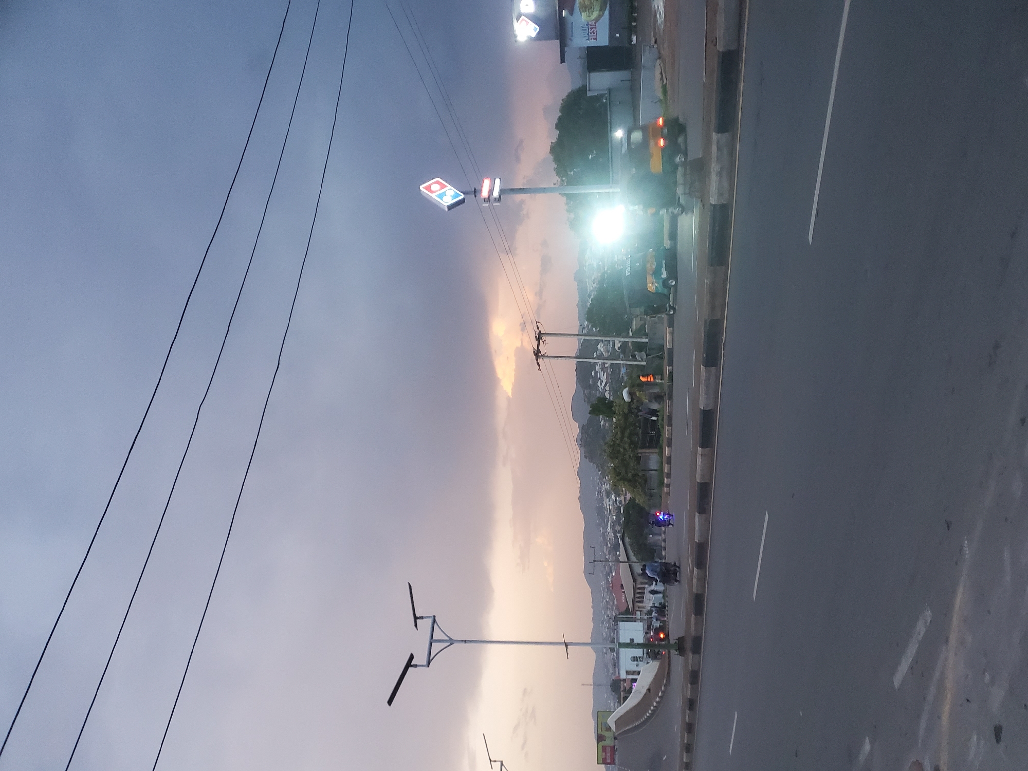 The image depicts a street scene in Jos, Nigeria, during dusk. The sky is overcast with a mix of dark clouds and areas of soft, warm light breaking through, suggesting sunset. Several power lines stretch across the upper portion of the frame.
In the foreground, a paved road with lane markings is visible. On the right side of the road, a prominent Domino's Pizza sign is illuminated, its iconic red and blue diamond logo clearly visible atop a pole. Next to it, another sign displays "NAIJA FIESTA."
Several vehicles are present. Two auto-rickshaws, often referred to as "Keke Napep" in Nigeria, are on the road. One is dark green and yellow, and the other is a lighter shade of yellow. They are illuminated by bright headlights, suggesting they are in motion. Two motorcycles are also on the road, one with a rider and the other appearing to be a solo rider with a blue light on the rear. A few people are visible in the mid-ground, some standing and others on motorcycles.
Further back, a street lamp with two solar panels is prominently featured on the left side of the road. Beyond the road, a cityscape with buildings and hills is visible. The overall impression is a busy urban intersection as the day transitions into evening.