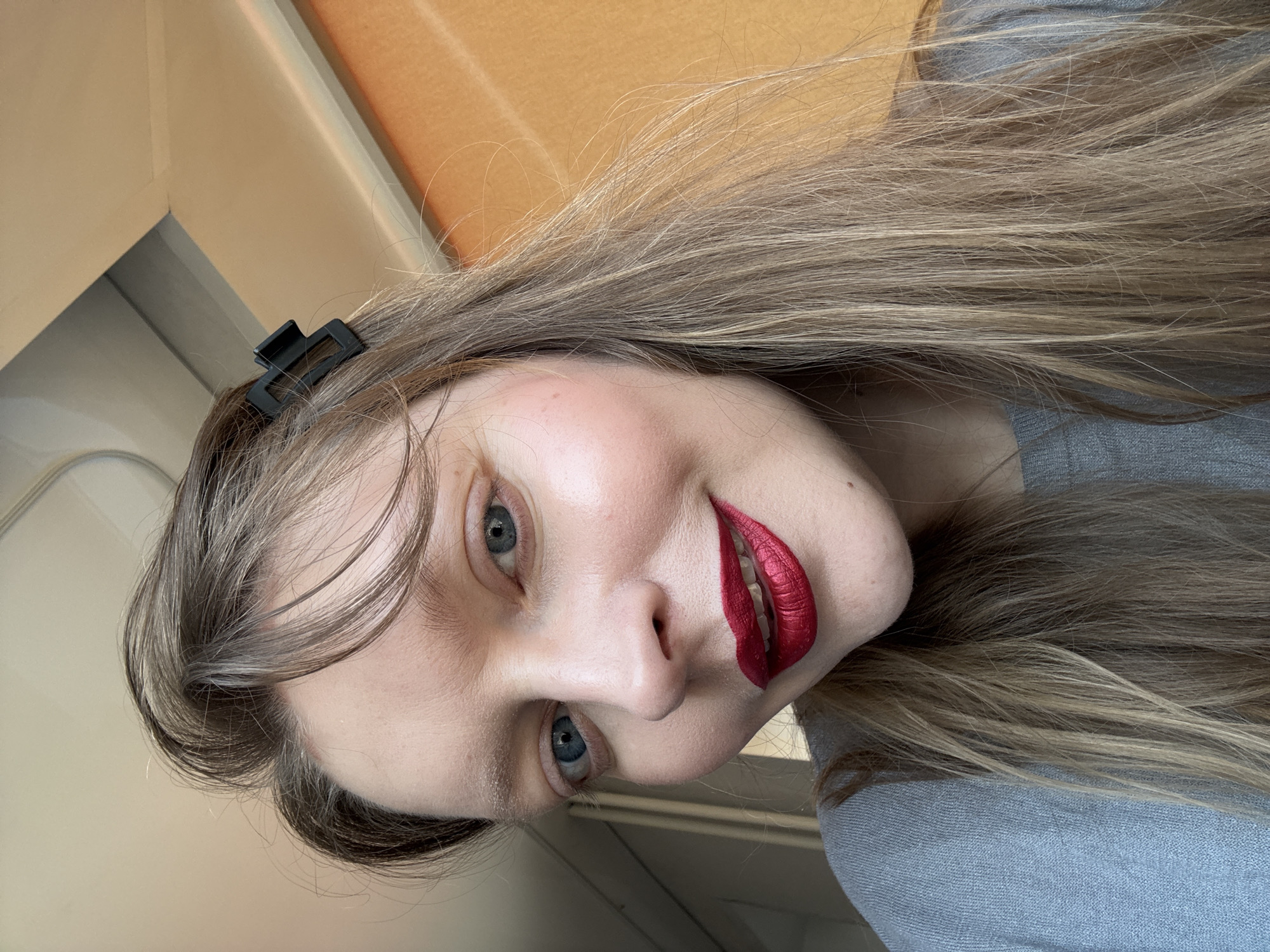 This is a close-up portrait of a young woman taken indoors. The woman is the primary subject, with her head tilted to the side and a slight, pleasant smile on her face. She has long, straight hair that is a mix of light brown and blonde highlights, appearing somewhat disheveled but natural. A black, rectangular hair clip is visible on the left side of her head, holding back some hair.
Her facial features are prominent, including fair skin, rosy cheeks, and bright blue eyes. A striking detail is her vibrant, metallic red lipstick. She is wearing a grey, textured garment, possibly a sweater or top. The background consists of plain, light beige and orange-brown walls or ceiling, indicating an indoor setting with simple architecture. The lighting is soft and even, illuminating her features. No text is visible in the image. The scene suggests a casual moment, possibly a selfie. The contextual location is Izhevsk, Russia.
