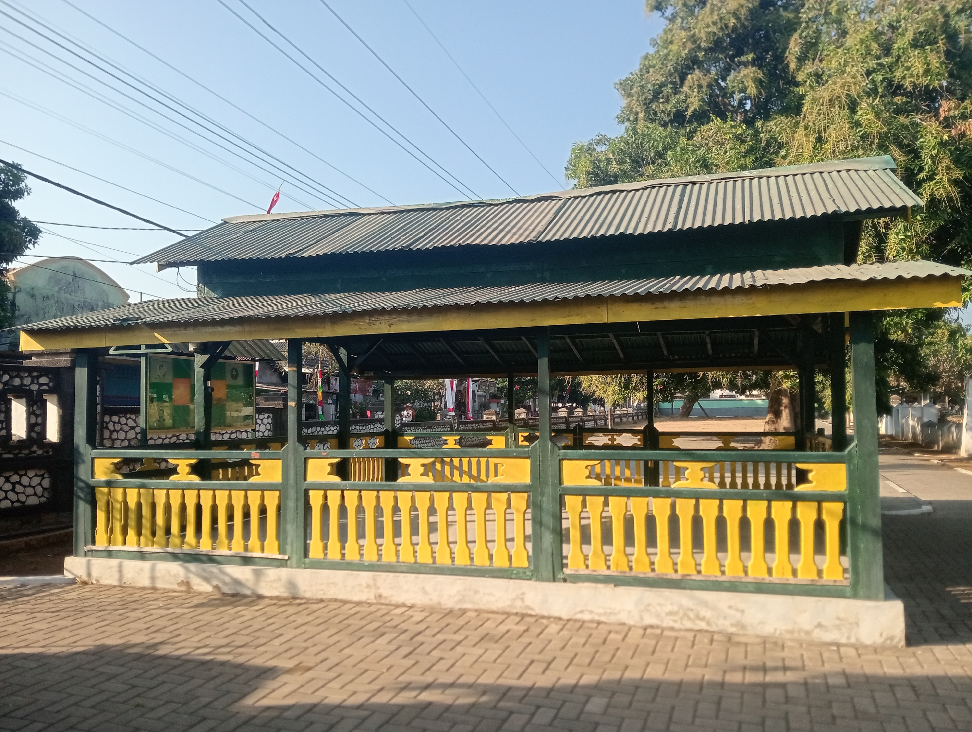The image depicts an outdoor covered pavilion with a corrugated metal roof, painted dark green and yellow. The structure is supported by green posts and features a yellow railing with decorative elements. The ground in front of the pavilion is paved with brick. In the background, there are trees, utility poles with numerous wires, and a hint of buildings. A small red and white flag is visible on the roof of the pavilion. The scene appears to be a public space, possibly a rest area or gathering point. The context provided indicates this location is in Bloro, Indonesia.
