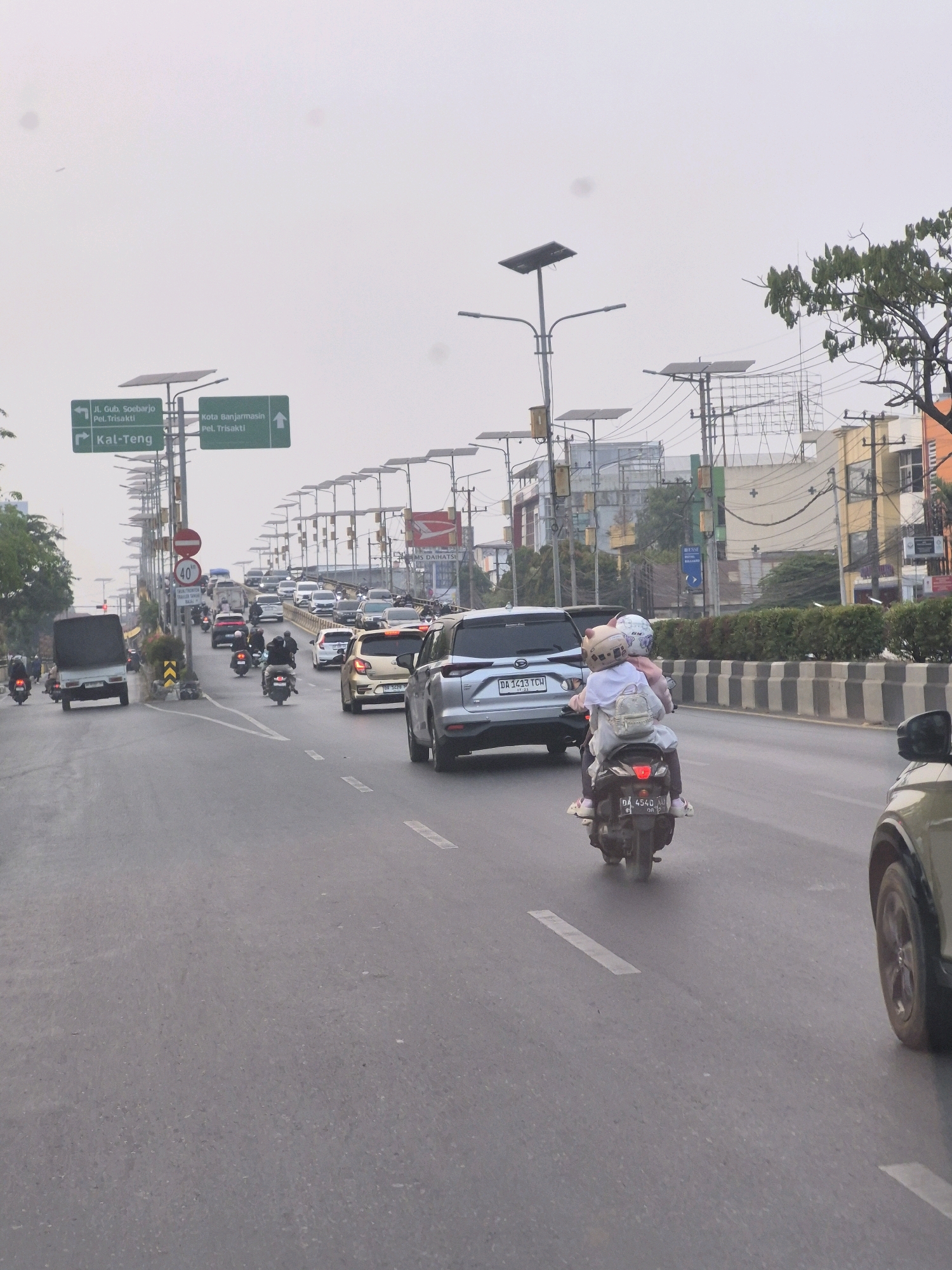 The image depicts a street scene in Banjarmasin, Indonesia, characterized by a moderate amount of traffic on an overcast day. The road appears to be a multi-lane highway or overpass.
In the foreground, a motorcycle with two passengers is moving away from the camera. The passenger in the rear is wearing a light-colored backpack and a helmet with cat-ear embellishments. The driver is wearing a helmet. The license plate of the motorcycle is partially visible, reading "DA 4540 AU". To the right of the motorcycle, a portion of a light-colored SUV is visible.
Further down the road, there is a flow of vehicles including cars, trucks, and motorcycles. A silver SUV with the license plate "DA 1433 TCH" is also visible. There are traffic signs indicating directions to "JL. GUB. SOEHARJO PEL. TRISAKTI" and "Kota Banjarmasin PEL. TRISAKTI". Another sign indicates "KAL-TENG". A speed limit sign showing "40" is also present.
The environment includes buildings with advertisements, streetlights equipped with solar panels, power lines, and some vegetation. The overall atmosphere is hazy, suggesting either a cloudy or polluted sky. The road surface is asphalt with painted lane markings. The presence of numerous traffic signals and infrastructure indicates a developed urban area.