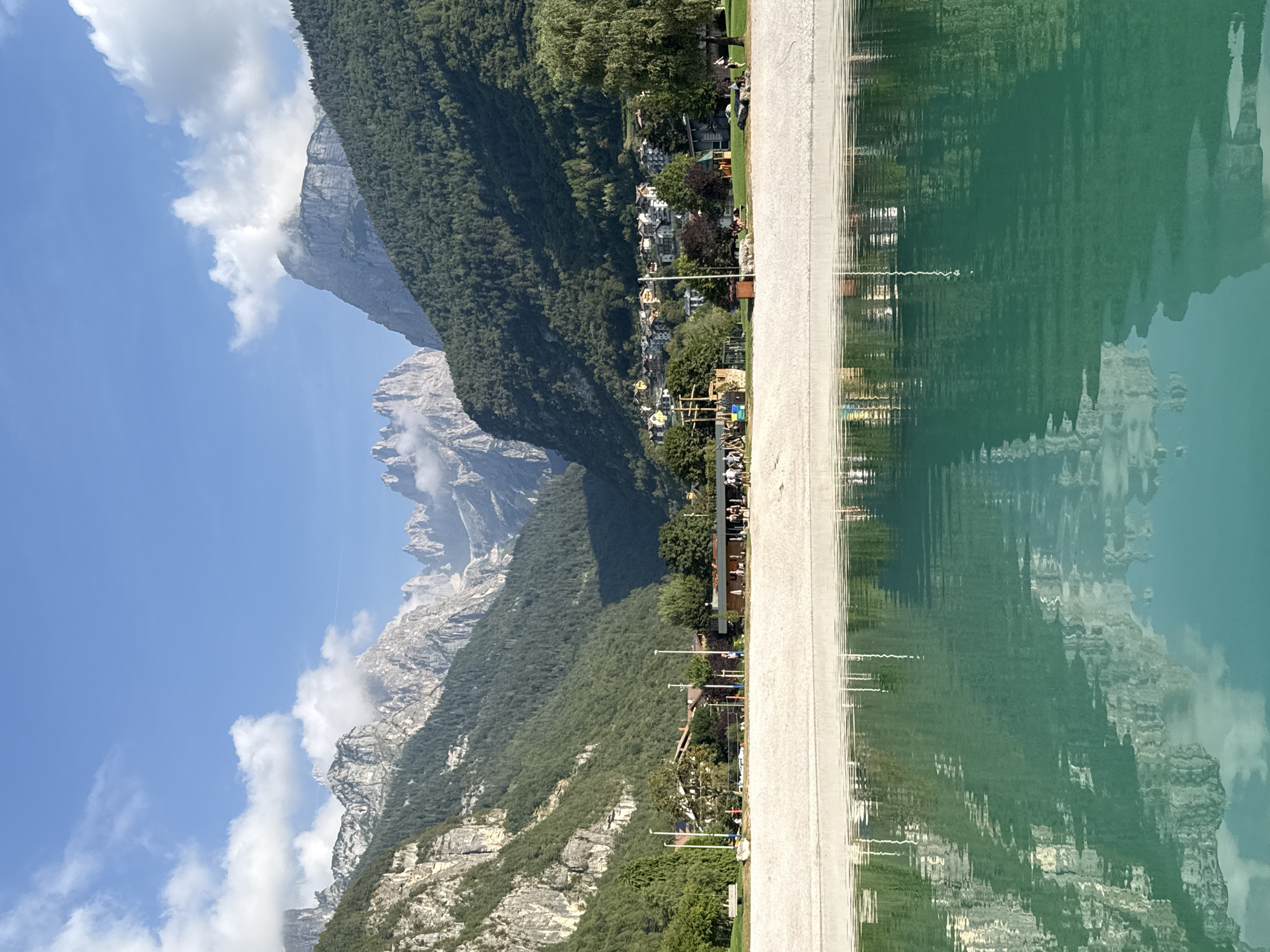 This is a horizontal, eye-level shot of a serene lake with a reflective surface, bordered by a sandy beach and a backdrop of lush green mountains. The water is a vibrant turquoise, reflecting the sky, clouds, mountains, and the buildings along the shore. The beach is smooth and light-colored, with a few scattered lounge chairs and umbrellas. Beyond the beach, a small town with red-tiled roofs is nestled at the foot of the mountains. The mountains themselves are majestic, with rugged peaks and dense forests covering their slopes. The sky is a clear blue, with a few scattered white clouds. The scene appears to be from the town of Molveno, Italy, renowned for its stunning lake and surrounding Dolomite mountains.