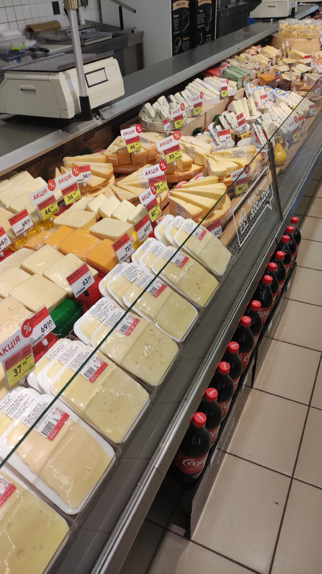 This is a top-down, medium shot of a cheese display in what appears to be a supermarket. The display is packed with various types of cheese, many of which are sliced and packaged. There are also some larger blocks and wedges of cheese visible. Red and white "АКЦІЯ" (meaning "SALE" in Ukrainian) labels with percentages are prominently displayed on many of the cheese packages.
To the right of the cheese display, there is a row of dark-colored bottles, likely soda, with red caps. The bottles have the Coca-Cola logo visible on them.
The setting is a well-lit retail environment, with metal shelving and glass barriers. The floor tiles are visible in the lower right corner of the image. Some signage is visible above the cheese display, including what appears to be an "8:00 to 22:00" time indication, suggesting operating hours. No people are visible in the frame. The overall impression is a commercial display of food products.