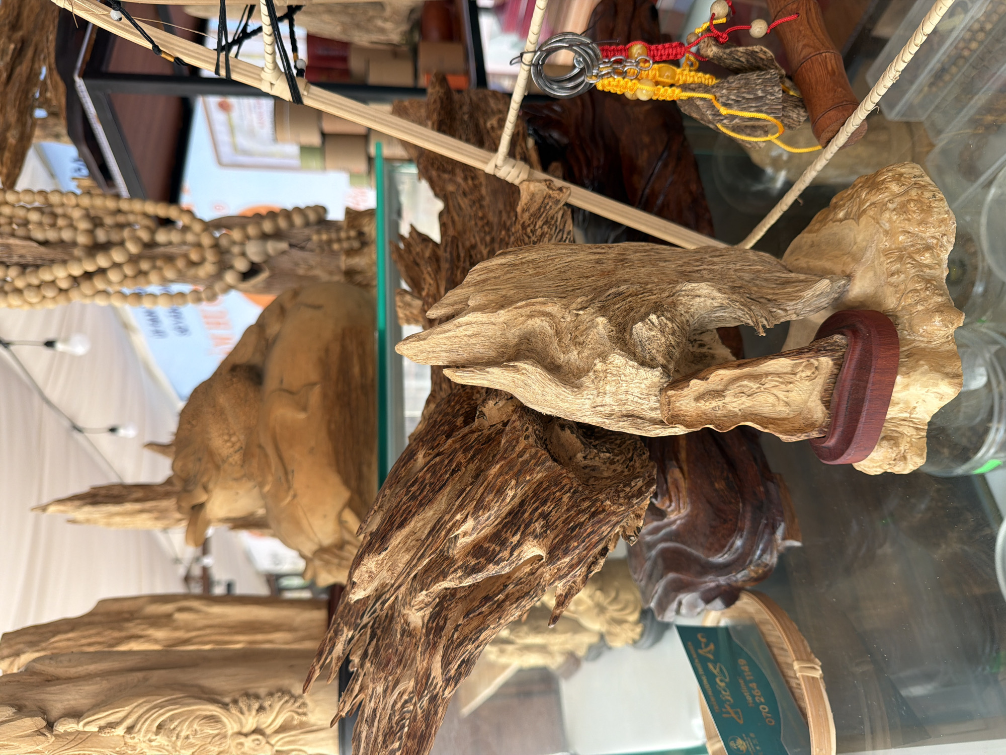 The image depicts a display of carved wooden objects, likely at a market or shop in Nha Trang, Vietnam. In the foreground, a piece of rough, textured wood is prominently displayed on a lighter, carved wooden base, which is itself on a glass shelf. To the left, a business card with Vietnamese text and a hotline number (070 264 1149) is visible on a small woven tray. Further back, other carved wooden items and strands of wooden beads are visible. The lighting suggests an indoor setting, possibly a stall at a tourist-oriented market. The overall impression is one of artisanal crafts made from natural wood.