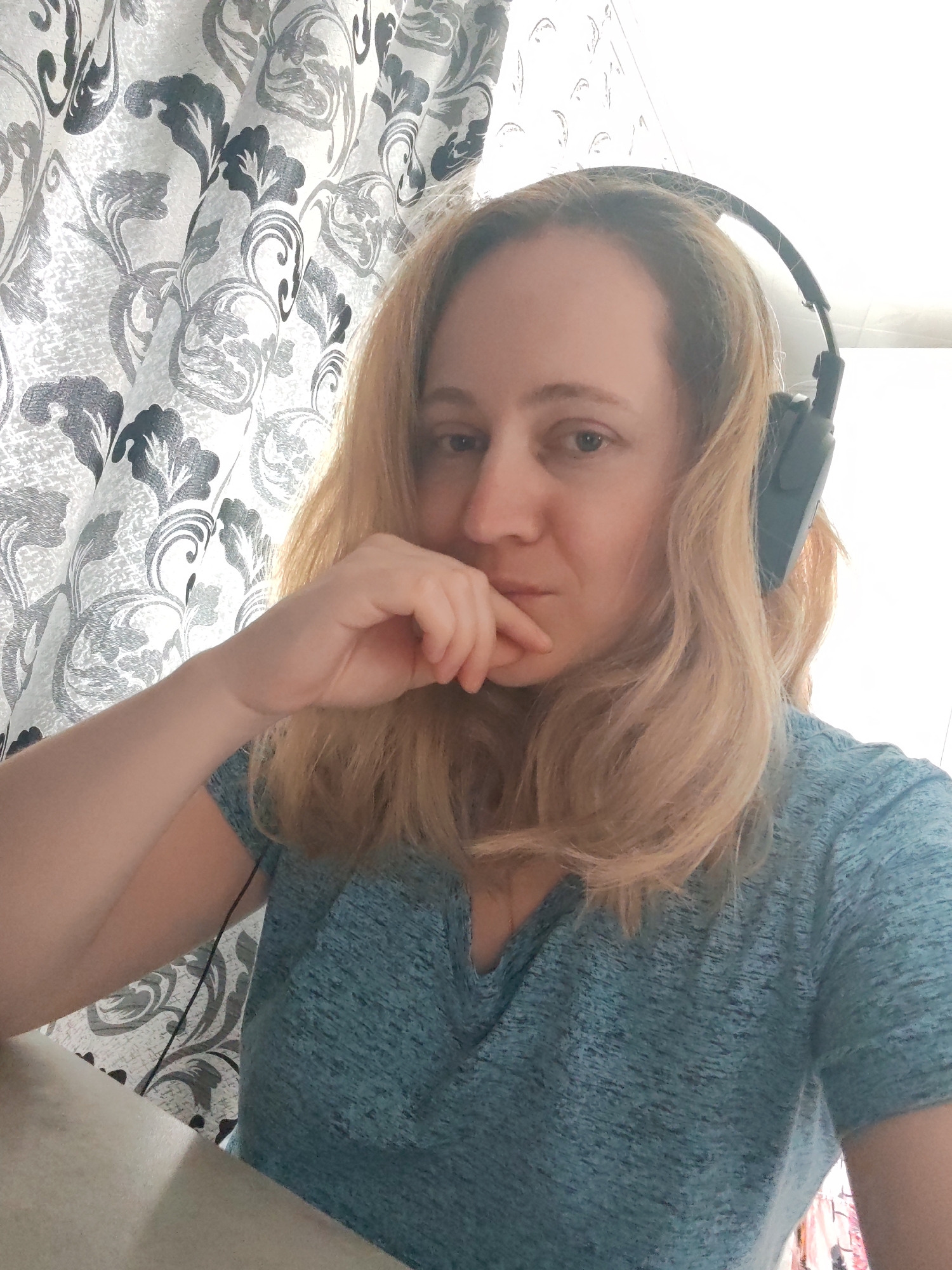 A woman with shoulder-length blonde hair, wearing a teal t-shirt and headphones, is shown in a selfie from the chest up. She is looking towards the camera with her right hand resting on her chin. The background consists of a patterned curtain with gray and black floral motifs. The woman is positioned in front of a desk or table, with a cable from the headphones visible. The scene appears to be indoors. The city of Сокол, Russia, is not depicted in the image.