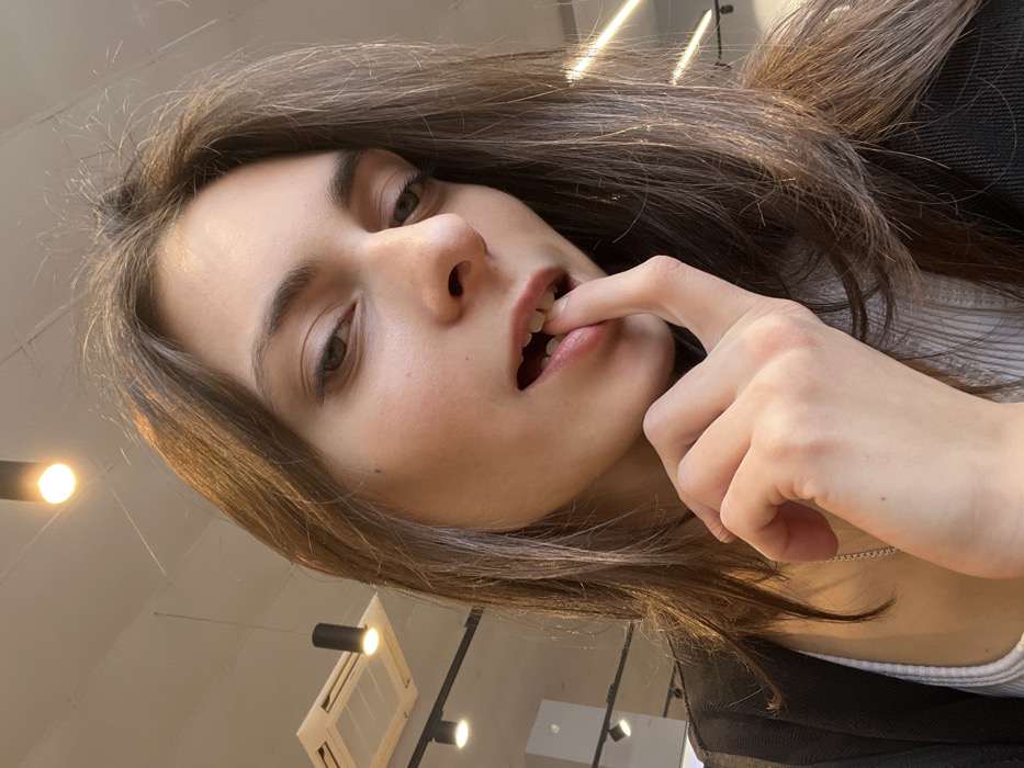A woman with brown hair and fair skin gazes into the camera. Her finger is placed inside her slightly parted lips, with a hint of lipstick visible. She is wearing a white top and a dark jacket. The background includes a ceiling with a light fixture, creating an indoor setting. The angle is slightly tilted, presenting a unique perspective.