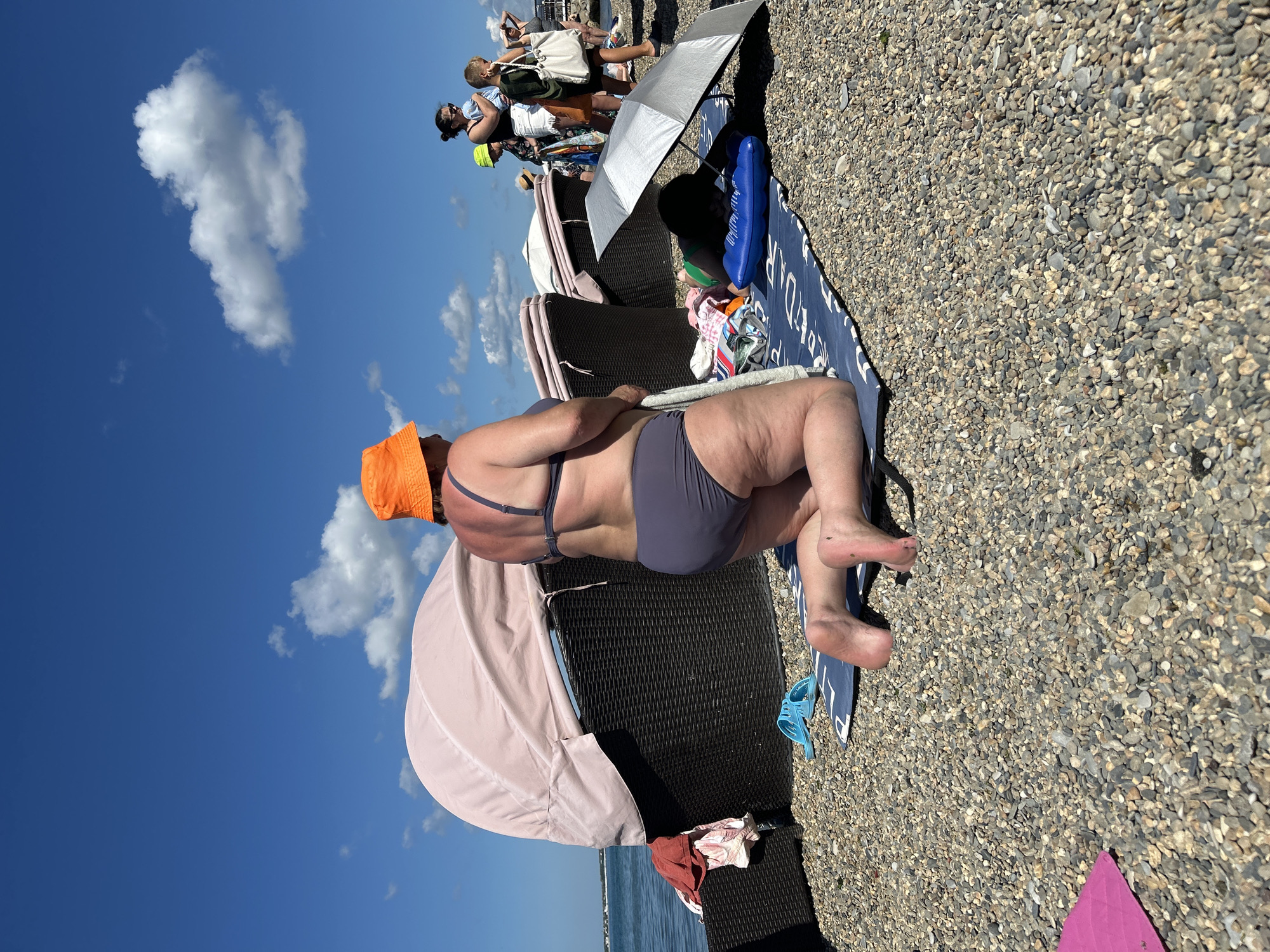In Gelendzhik, Russia, on a bright, sunny day with a blue sky and scattered clouds, a woman kneels on a pebble beach next to the sea. She is wearing a grey bikini and an orange bucket hat. In front of her is a blue and white beach towel, and to her left are several pieces of clothing. Behind her are dark wicker chairs with pink canopy coverings. To the right, another person lies under a silver umbrella on a blue inflatable. Further in the background, other beachgoers are visible, some with children.