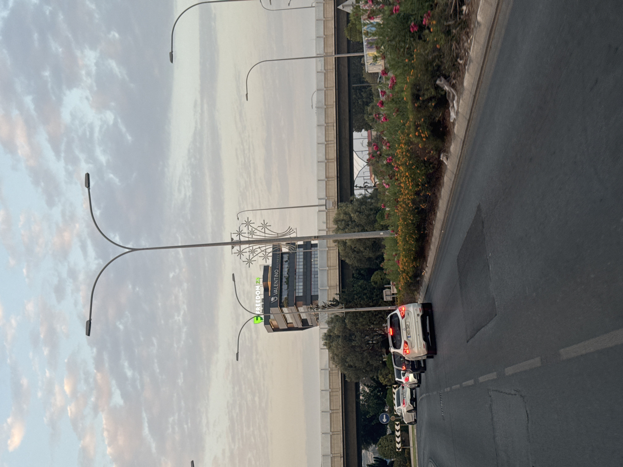 The image captures a view of a street in Germasogeia Municipality, Cyprus, with several cars driving on a road. In the background, a modern building with "FREEDOM 24" and "VALENTINO BUSINESS CENTER" signage is visible. A highway overpass runs across the scene, supported by concrete pillars. The sky is overcast with clouds, and there is a median strip with flowering plants and trees. The road has visible lane markings and some patches. The overall atmosphere suggests an urban setting, possibly during the day or early evening due to the lighting.