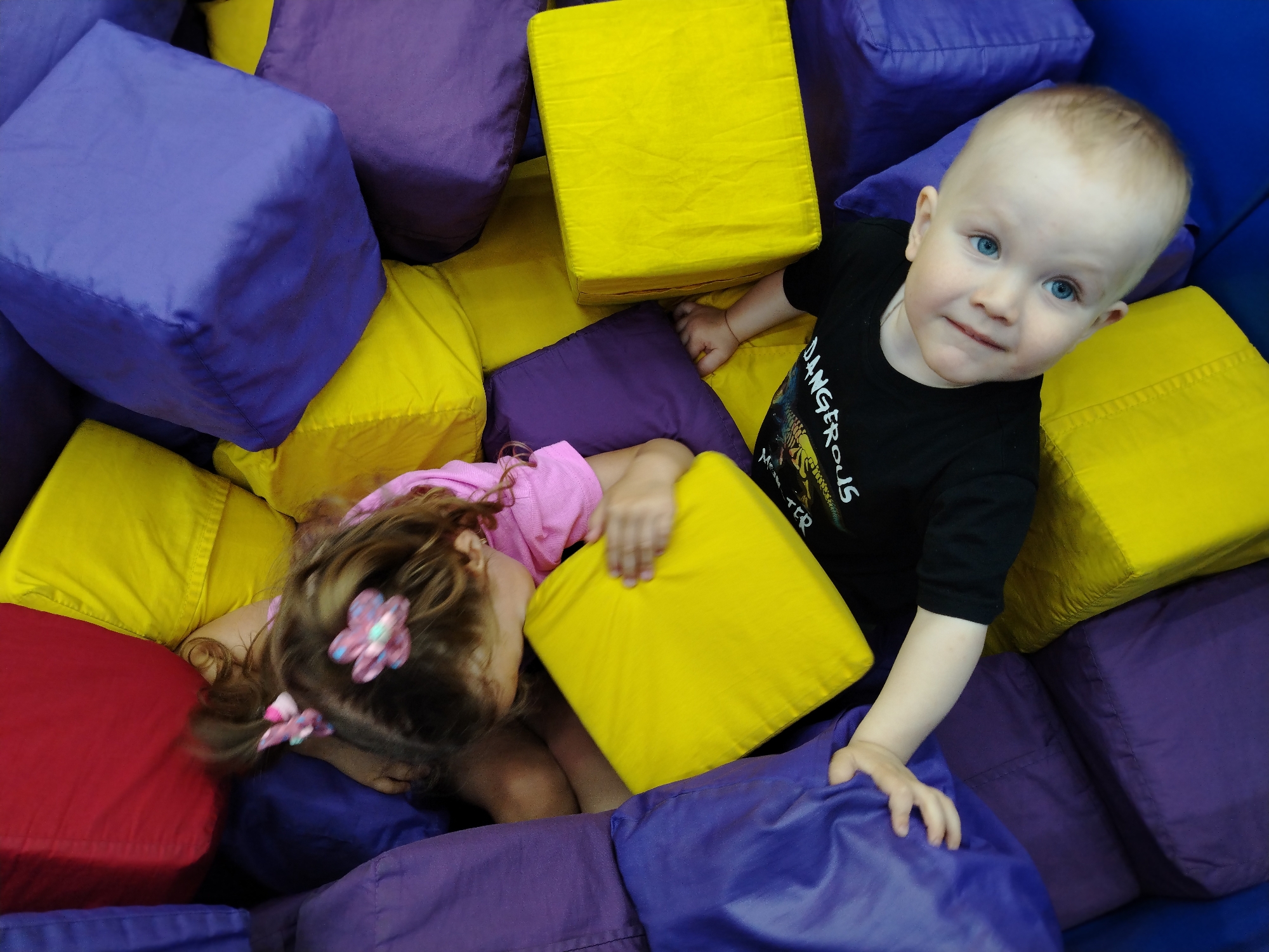 The image captures two young children within a soft play environment filled with numerous large, fabric-covered, cube-shaped blocks. The dominant colors of these blocks are bright yellow and dark purple/blue, with one red block visible in the lower-left portion of the frame.
In the upper-right quadrant, a male toddler, estimated to be between 1 and 3 years old, is seated or leaning among the blocks. He possesses fair skin, light-colored hair, and distinct blue eyes, maintaining direct eye contact with the camera. He is wearing a black short-sleeved t-shirt featuring a white graphic print on the chest, with the word "DANGEROUS" partially legible. His right arm is visible, with his hand resting on a yellow block.
In the lower-left quadrant, a female child, estimated to be between 2 and 5 years old, is mostly obscured by the blocks, appearing to be lying or immersed within the padded structure. Her medium brown, wavy hair is visible, adorned with two pink flower-shaped hair clips. She is wearing a pink t-shirt. Her head is turned away from the camera, facing downwards or towards the lower-left corner of the frame.
The children are in close proximity within the play area but are not directly interacting with each other at the moment the image was captured. Both are physically engaged with the soft blocks, which form the entirety of the visible environment. The specific location within Chaikovsky, Russia, cannot be ascertained from the visual evidence presented.