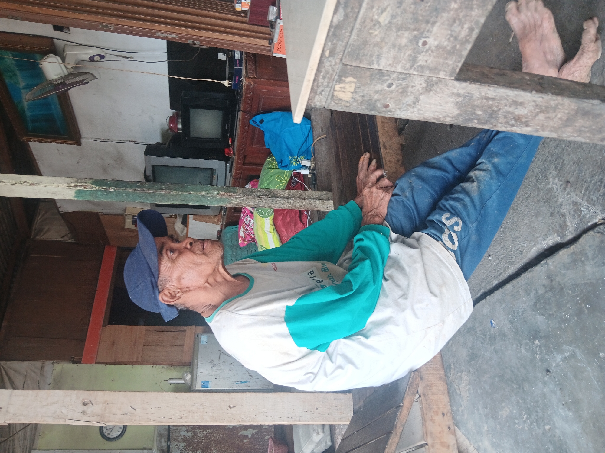 In Batursari, Indonesia, an elderly man wearing a blue baseball cap and a long-sleeved shirt with white and teal coloring sits on a wooden platform. The man's legs are crossed, and his hands rest on his lap. He is wearing blue pants. The background of the image reveals a cluttered interior space, with various objects such as televisions, a fan mounted on the wall, pillows, and what appears to be a refrigerator. Wooden beams and panels suggest a rustic construction. A wooden table is visible in the foreground. The overall impression is one of a simple, lived-in environment.