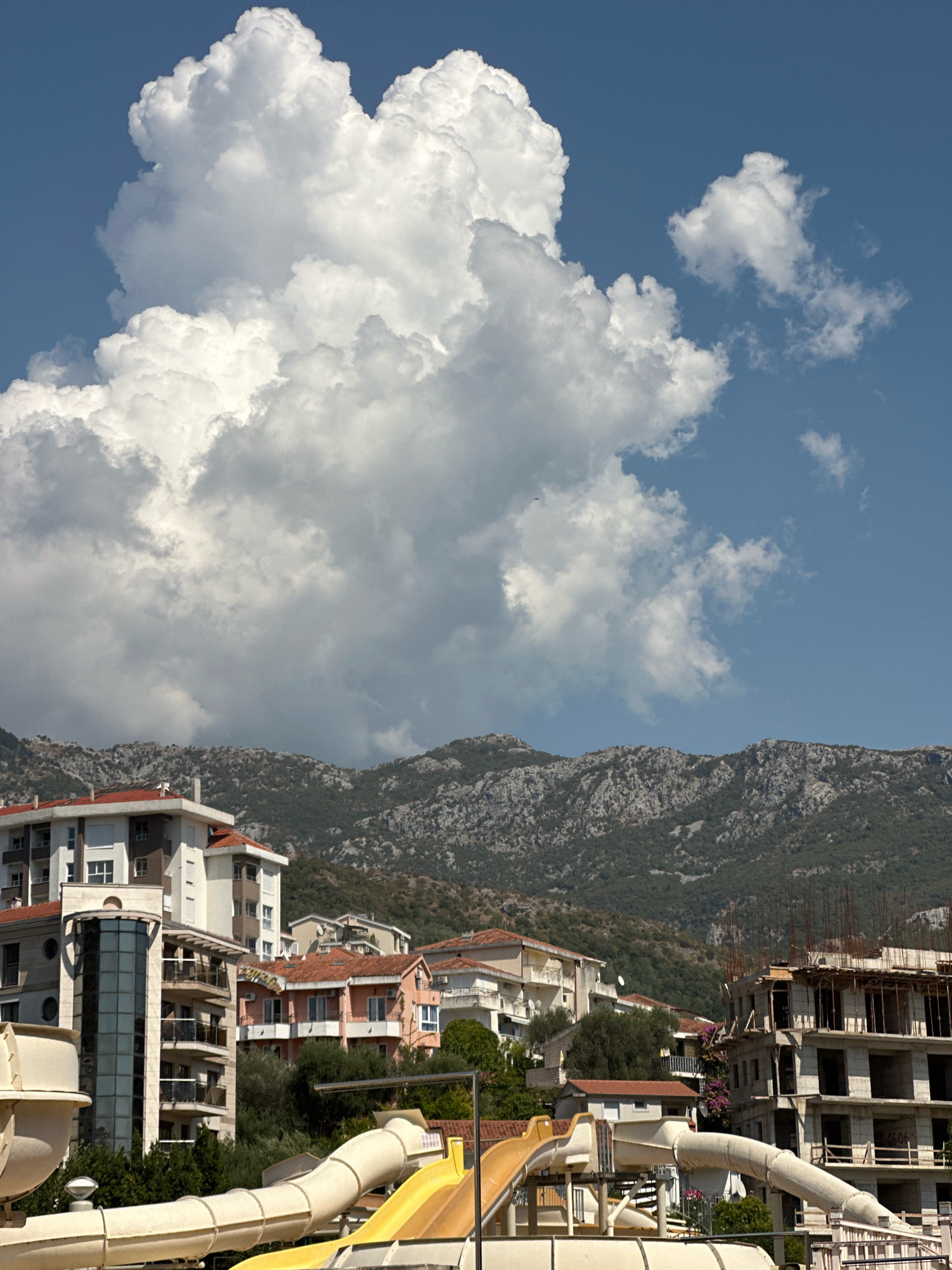 This is a medium-shot photograph taken outdoors, looking up at a clear blue sky dominated by a massive, billowy cumulus cloud formation. Below the sky and cloud, a mountainous landscape forms the backdrop, with rocky, barren peaks sparsely covered in vegetation. In the foreground, a collection of buildings and water slides are visible, suggesting a resort or residential area.
The buildings are varied in style and construction. On the left, a modern, multi-story building features a curved glass-enclosed elevator shaft and balconies. Adjacent to it, a building with a red tile roof and multiple windows is visible. Further to the right, another building with a red tile roof has balconies and several floors. To the far right, a multi-story structure appears to be under construction, with exposed rebar and concrete floors.
In the lower portion of the image, parts of a water park are depicted, with white and yellow water slides curving and snaking through the scene. Green foliage and trees are interspersed among the buildings and slides. The overall impression is a sunny day in a coastal town, with a mix of leisure facilities and ongoing development set against a natural mountainous backdrop. The specific location is Bečići, Montenegro.