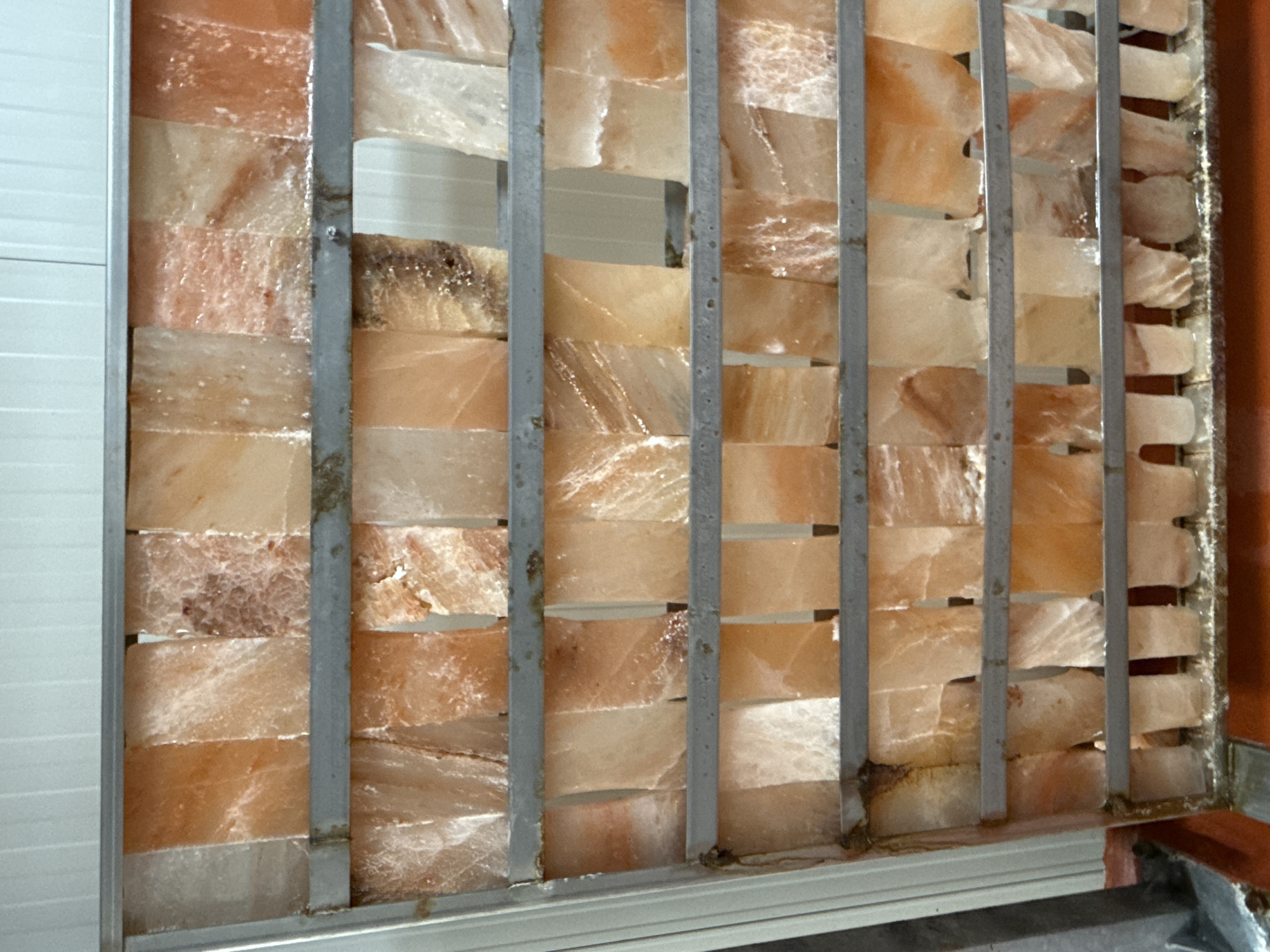 The image displays a close-up of an architectural or decorative partition constructed from numerous translucent blocks. These blocks vary in color from pale pink and off-white to warmer orange and reddish-brown hues, exhibiting characteristic crystalline patterns and veins, indicating they are likely Himalayan salt blocks. They are arranged in horizontal rows and held in place by several vertical, slender grey metal bars that run through them. Some of the blocks have irregular edges or show natural variations in texture and shape.
To the left of the salt block structure, a light-colored wall with distinct horizontal ridges or panels is visible. On the right side, a darker, reddish-brown element, possibly a wall section or frame, can be seen. The bottom of the structure includes a metallic frame or base. The blocks appear somewhat illuminated, emphasizing their translucent quality.
There are no people, visible activities, or events taking place in the image; it captures a stationary structure. No text is visible anywhere in the frame. The setting appears to be an interior space, likely in Stara Zagora, Bulgaria, possibly a salt room, spa, or a modern building incorporating natural materials.