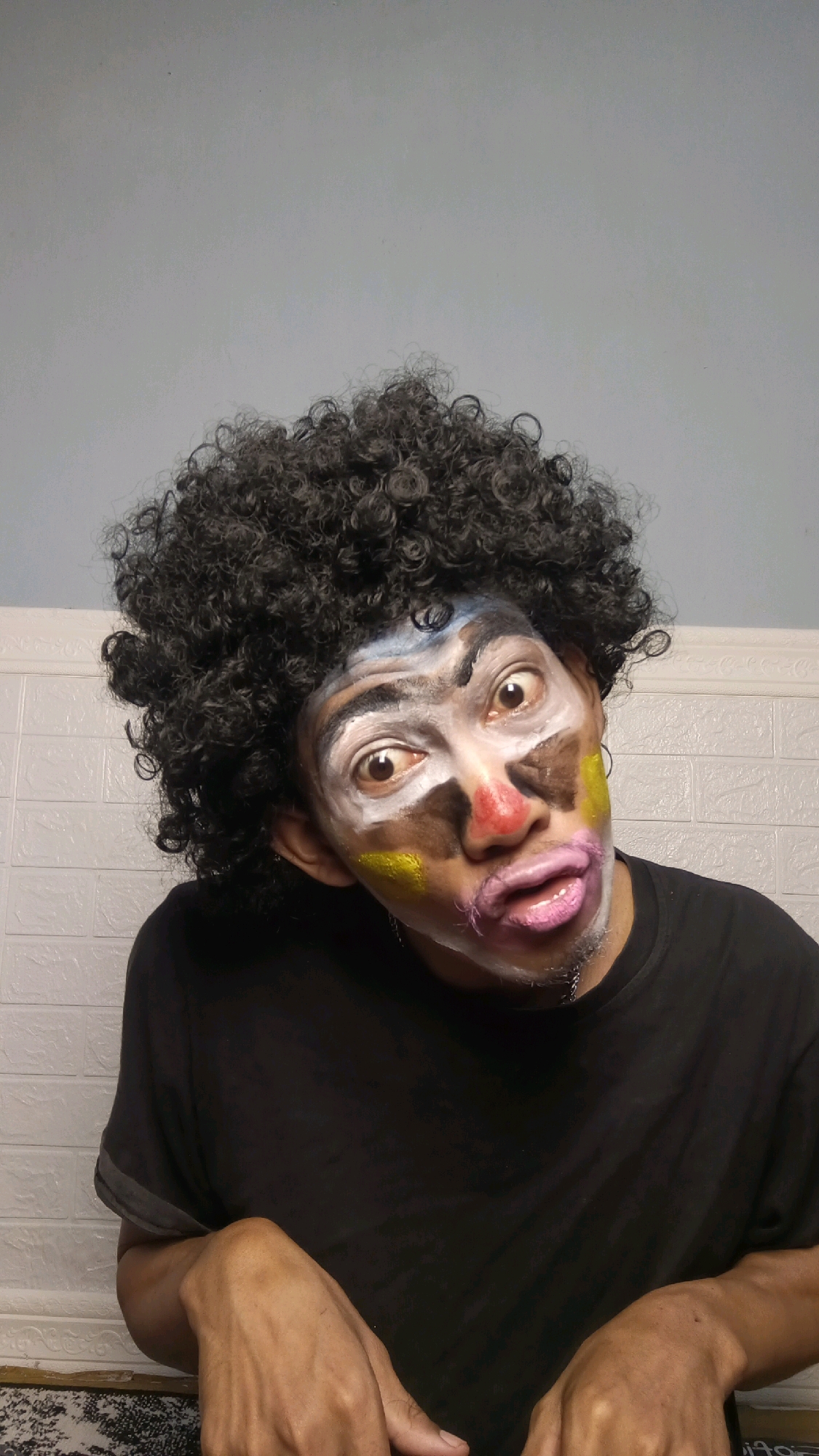 A person with curly black hair is wearing clown makeup and a black t-shirt. The makeup includes a white base, brown and blue accents on the face, a red nose, yellow markings on the cheeks, and pink lips. The person's eyes are wide open, and their head is tilted. They are indoors, with a white tiled wall in the background and a light blue wall above it. The lower portion of the image shows the person's hands resting on what appears to be a patterned surface. The location is described as Kebumen, Indonesia.