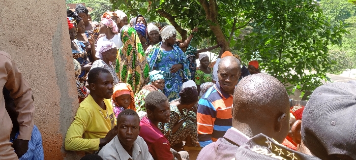 A group of people gathers outdoors, primarily Black individuals of varying ages, dressed in casual clothing and colorful, patterned fabrics. Some wear head coverings, like turbans and caps. The crowd is dense, with some individuals seated and others standing. To the left, a light brown wall is visible, and green trees fill the background. The scene seems to capture a community event or gathering.