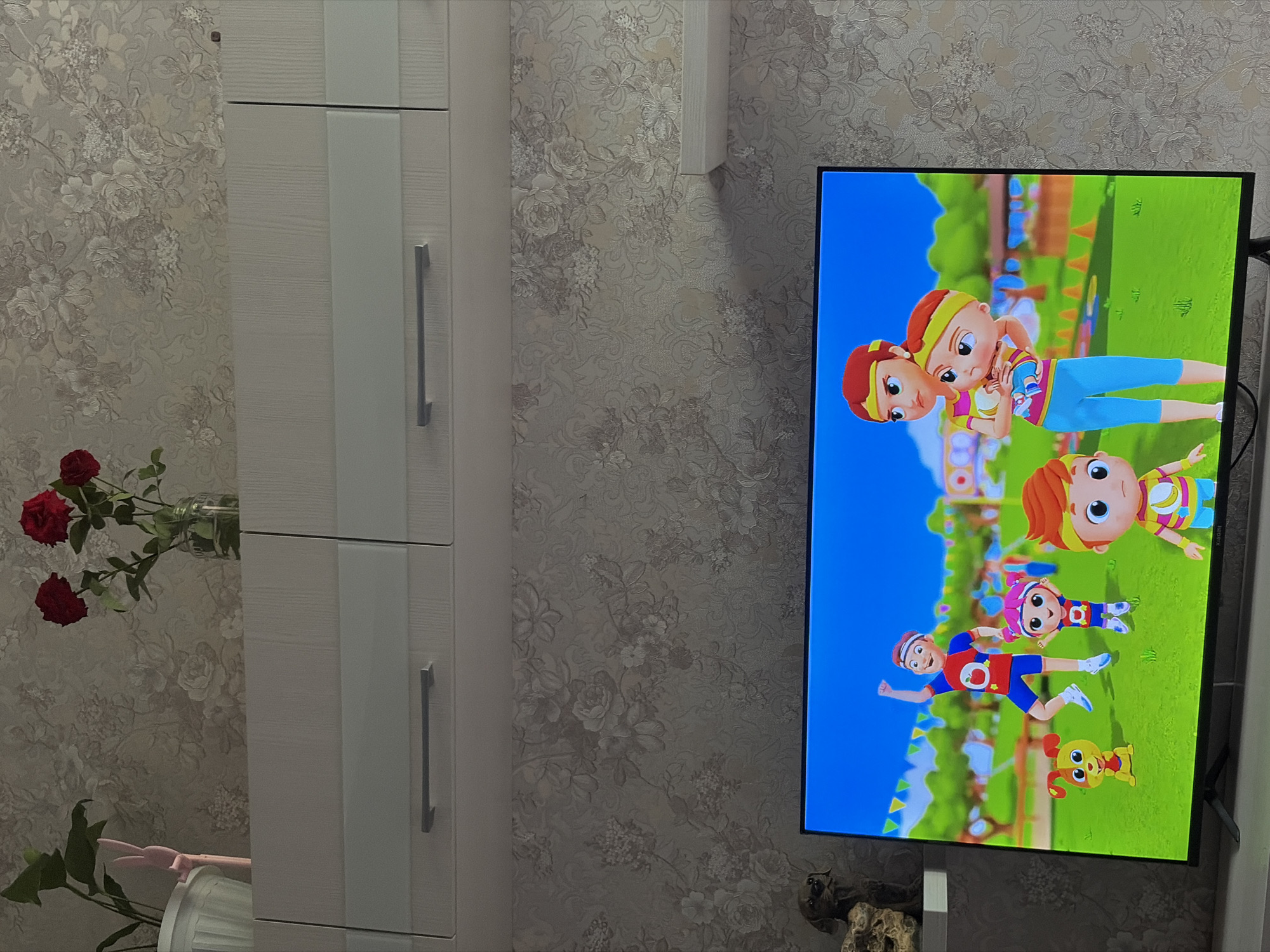 A television screen displays a cartoon scene with animated characters, including a male and female figure holding a baby, and other child-like characters in athletic attire, on a grassy field with a blue sky and decorative flags. To the left of the television, a shelf holds a statue of a dog and a plant. Above the television, a wall is adorned with floral wallpaper. A set of white cabinets is mounted on the wall above the television. On top of the cabinets, a vase holds red roses and a potted plant. The image was taken in Izhevsk, Russia.
